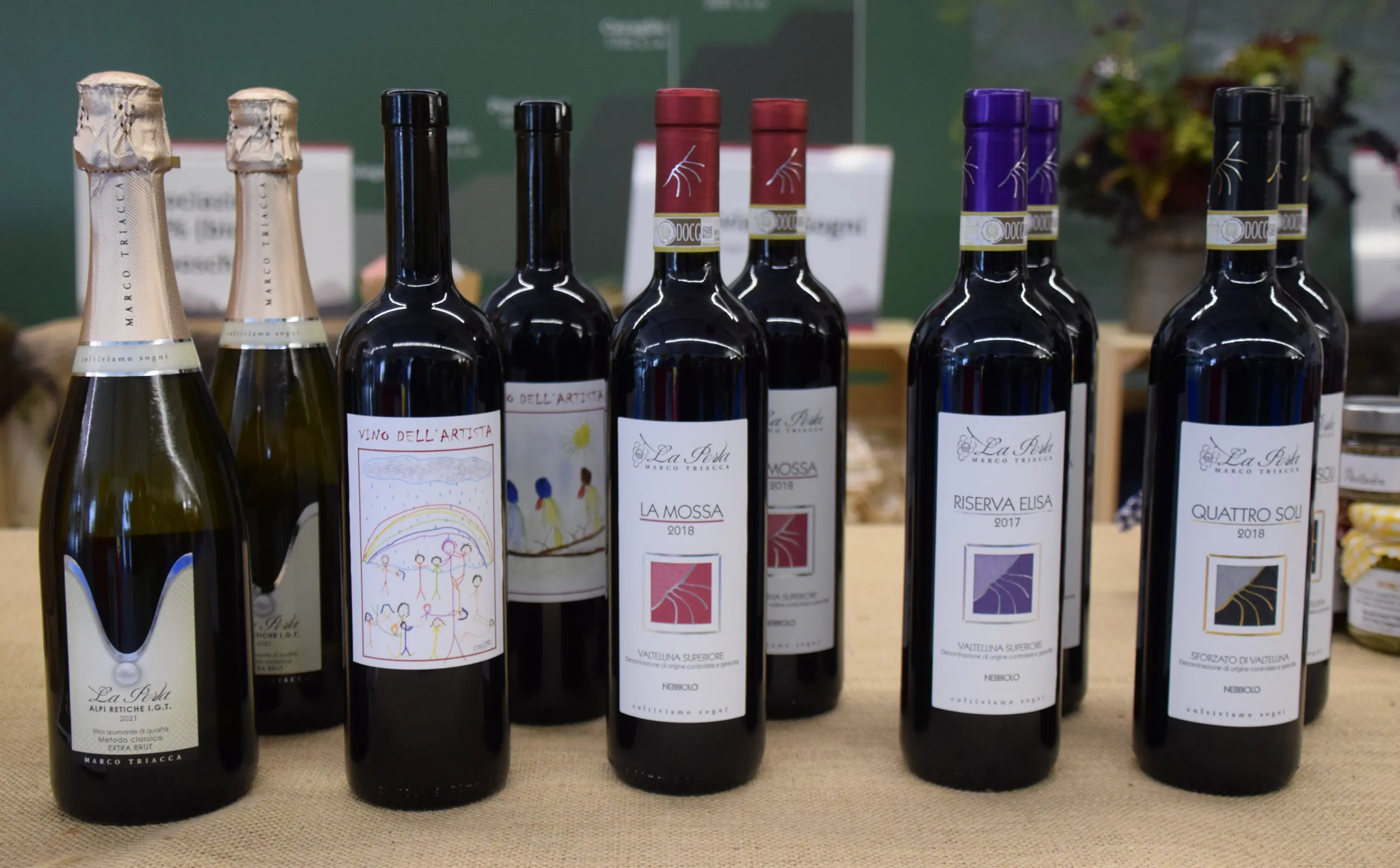 Vari tipi di vino in bottiglia su un tavolo. Le etichette mostrano diverse annate e nomi come "La Mossa", "Riserva Elisa" e "Quattro Sole". Il vino spumante è in primo piano a sinistra.