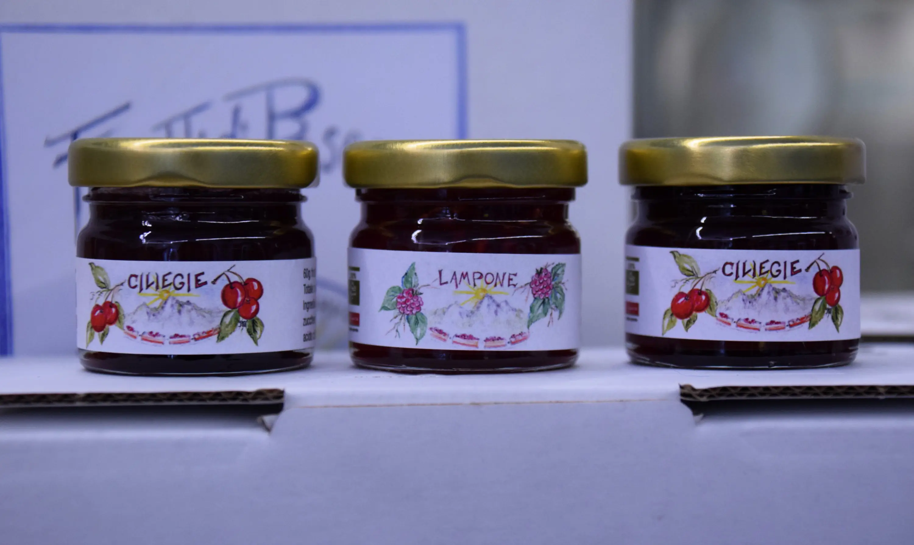 Tre barattoli di marmellata su uno sfondo bianco. I barattoli contengono marmellate di ciliegie, lamponi e ciliegie, ognuno con un'etichetta decorativa. I coperchi sono dorati.