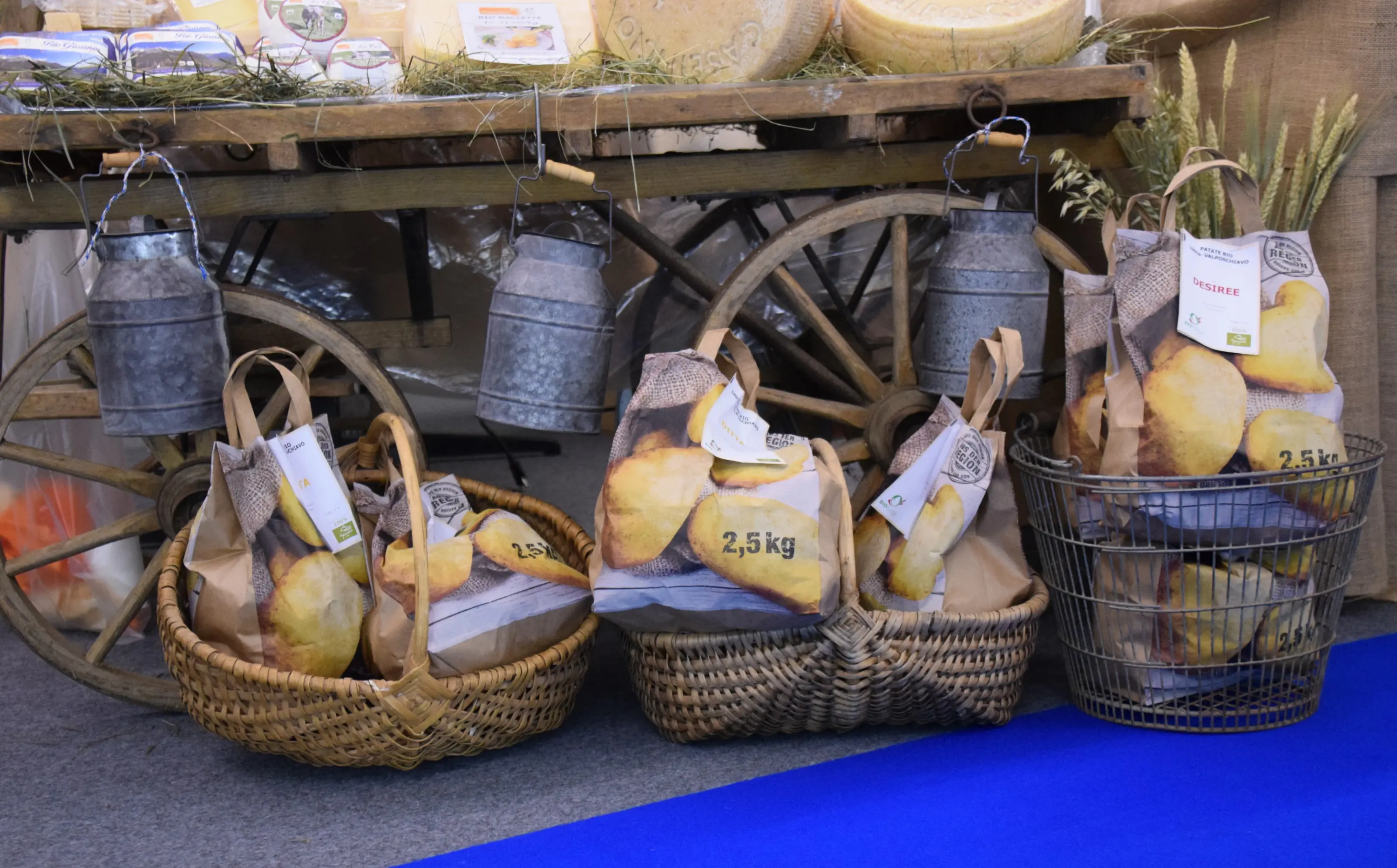 Cesti di paglia contenenti sacchi di patate, con etichette che indicano il peso. Sullo sfondo, un tavolo rustico e contenitori metallici.