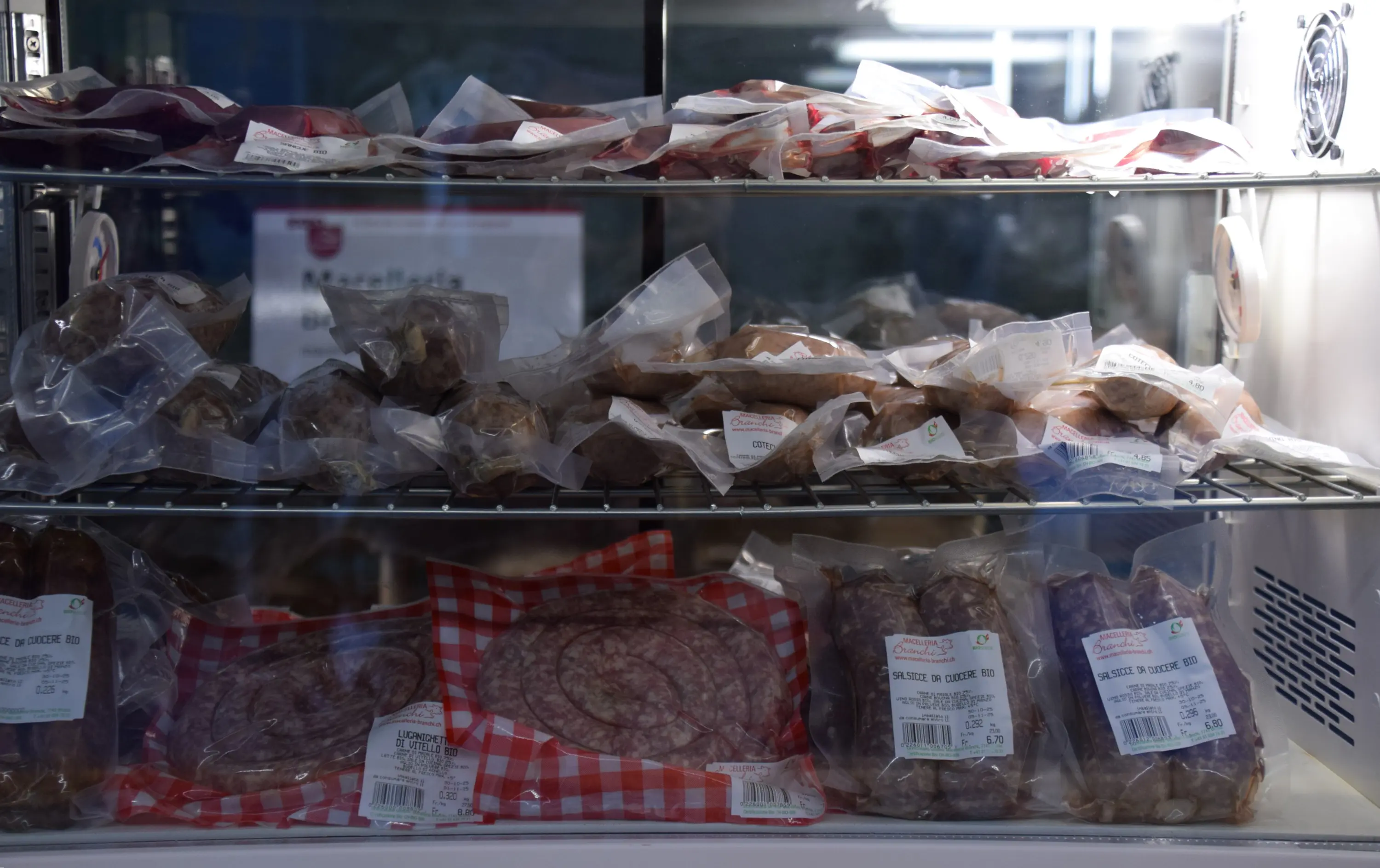 Vari tipi di salumi confezionati, disposti su ripiani di un frigorifero. I pacchetti presentano etichette informative e sono disposti ordinatamente.