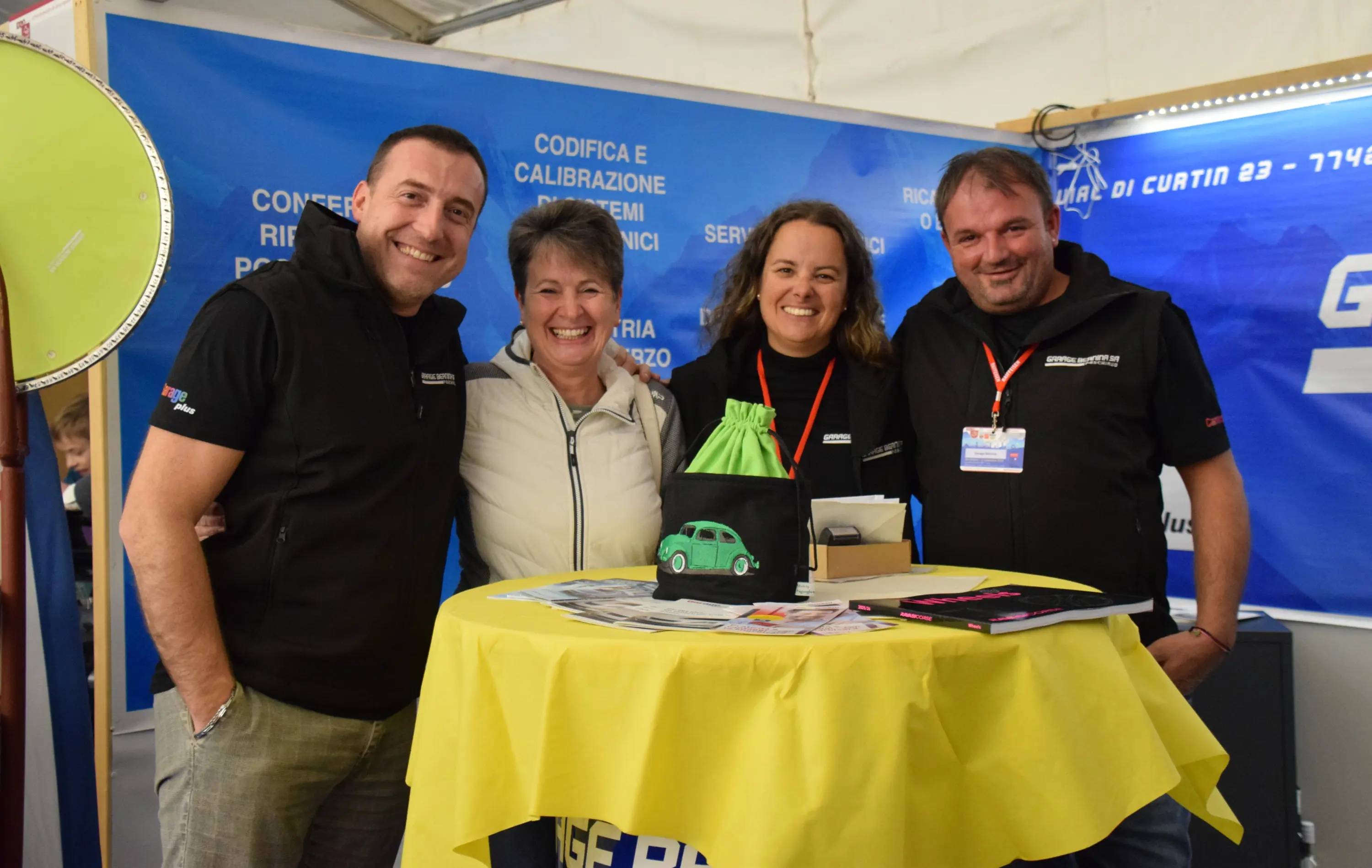 Quattro persone sorridenti posano insieme dietro a un tavolo giallo, allestito con materiali promozionali e un piccolo modello di auto, in un ambiente di fiera.