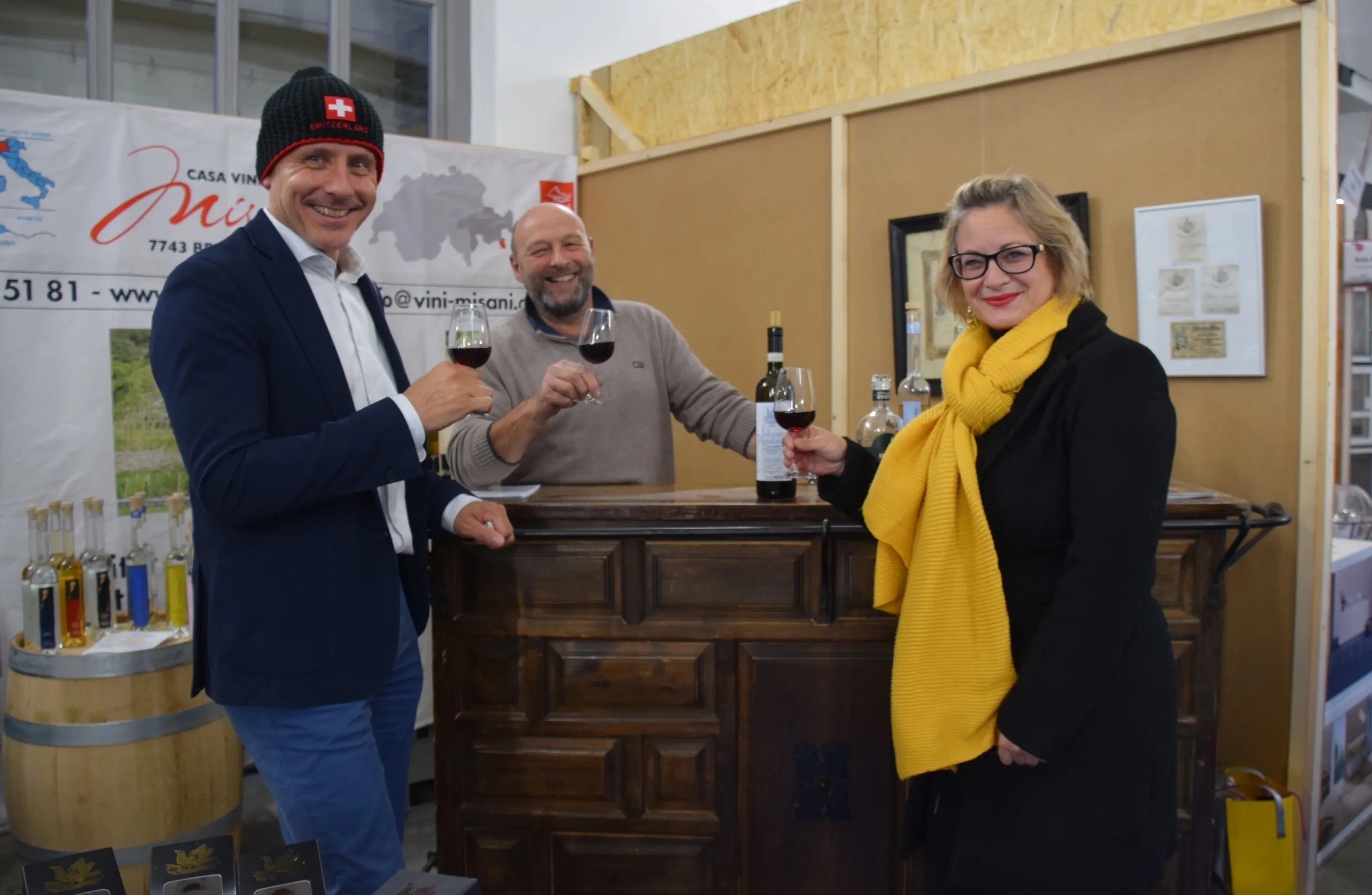 Tre persone brindano con bicchieri di vino presso un bancone in un evento di degustazione. Sullo sfondo ci sono bottiglie e un poster con la mappa della Svizzera. Un uomo indossa un berretto con la croce svizzera. La donna a des