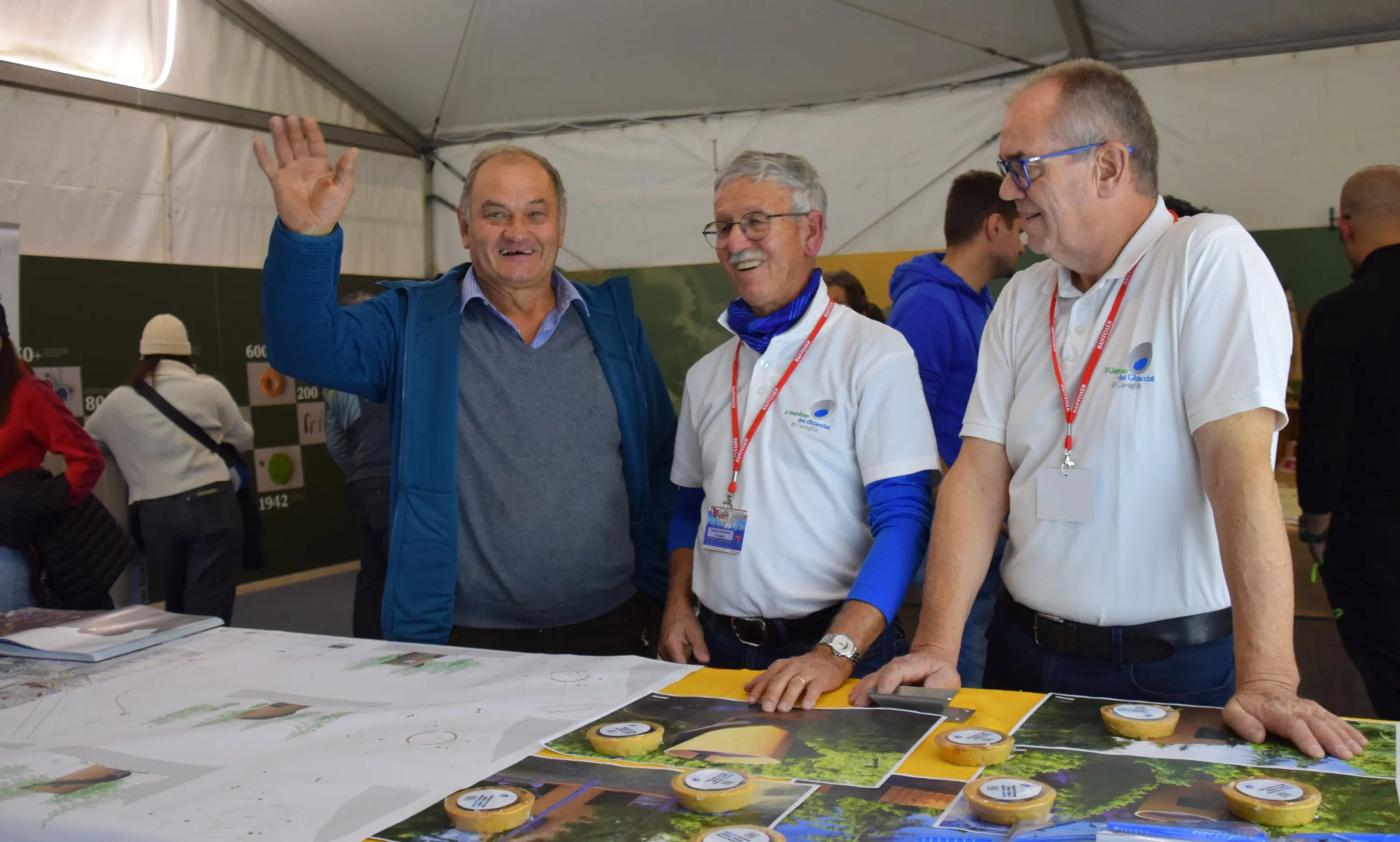 Tre uomini sorridenti all'interno di una tenda. Uno di loro saluta con la mano mentre gli altri due osservano un tavolo con dei materiali visivi e mappe. Tutti indossano magliette bianche con un logo.