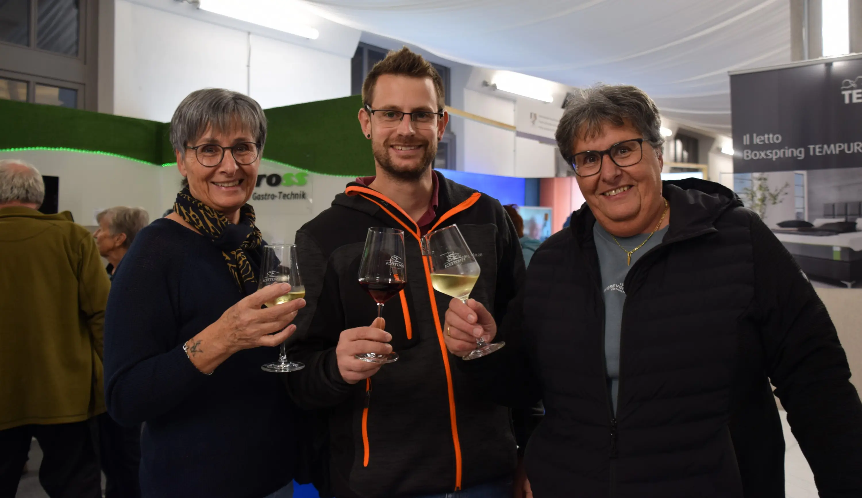 Tre persone sorridenti brindano con calici di vino, ognuna con una bevanda diversa, in un ambiente festoso.
