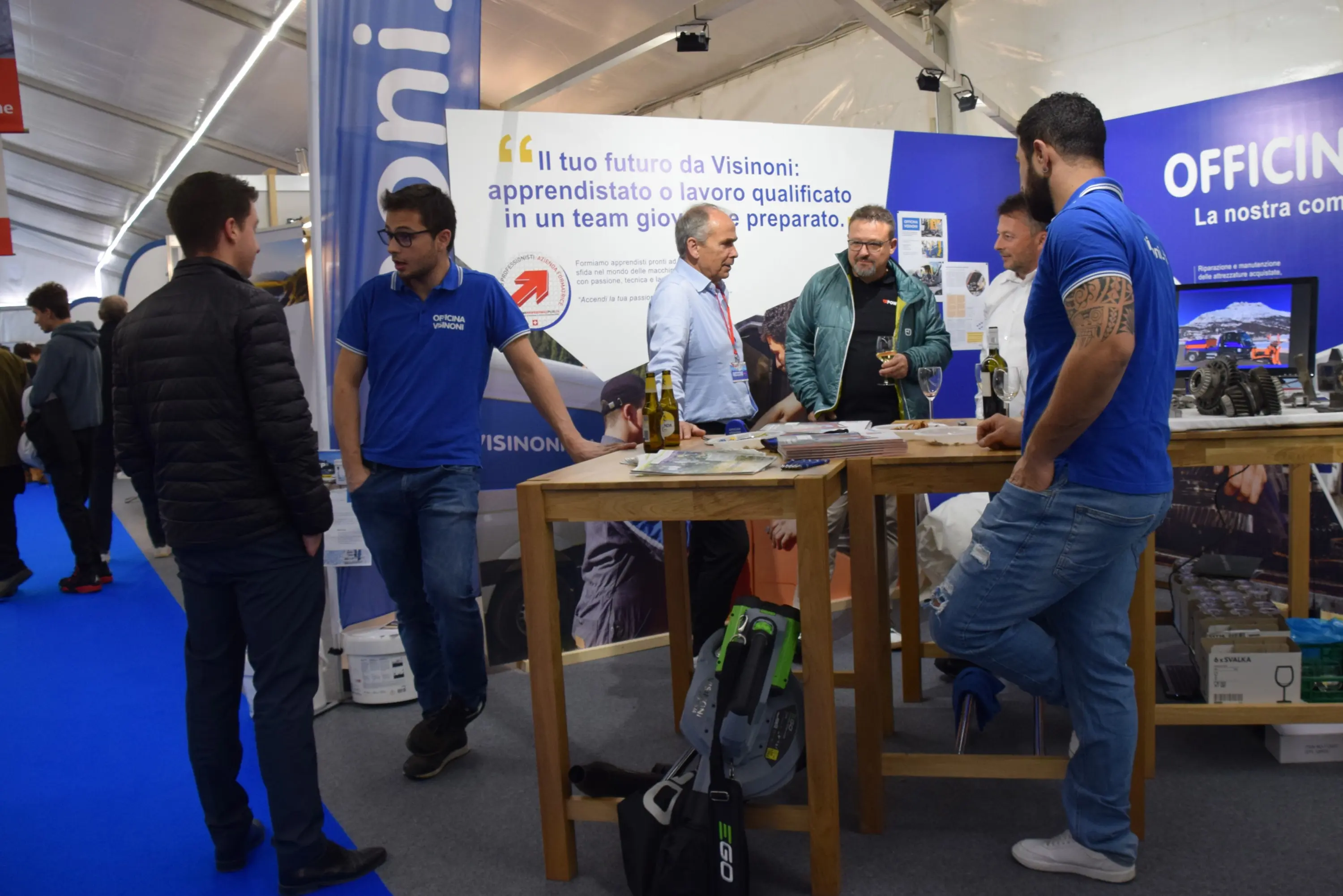 Un gruppo di persone interagisce in uno stand espositivo. Due uomini conversano con un rappresentante mentre altri sono riuniti attorno a un tavolo. Sfondi blu e grafiche promozionali di un'azienda.