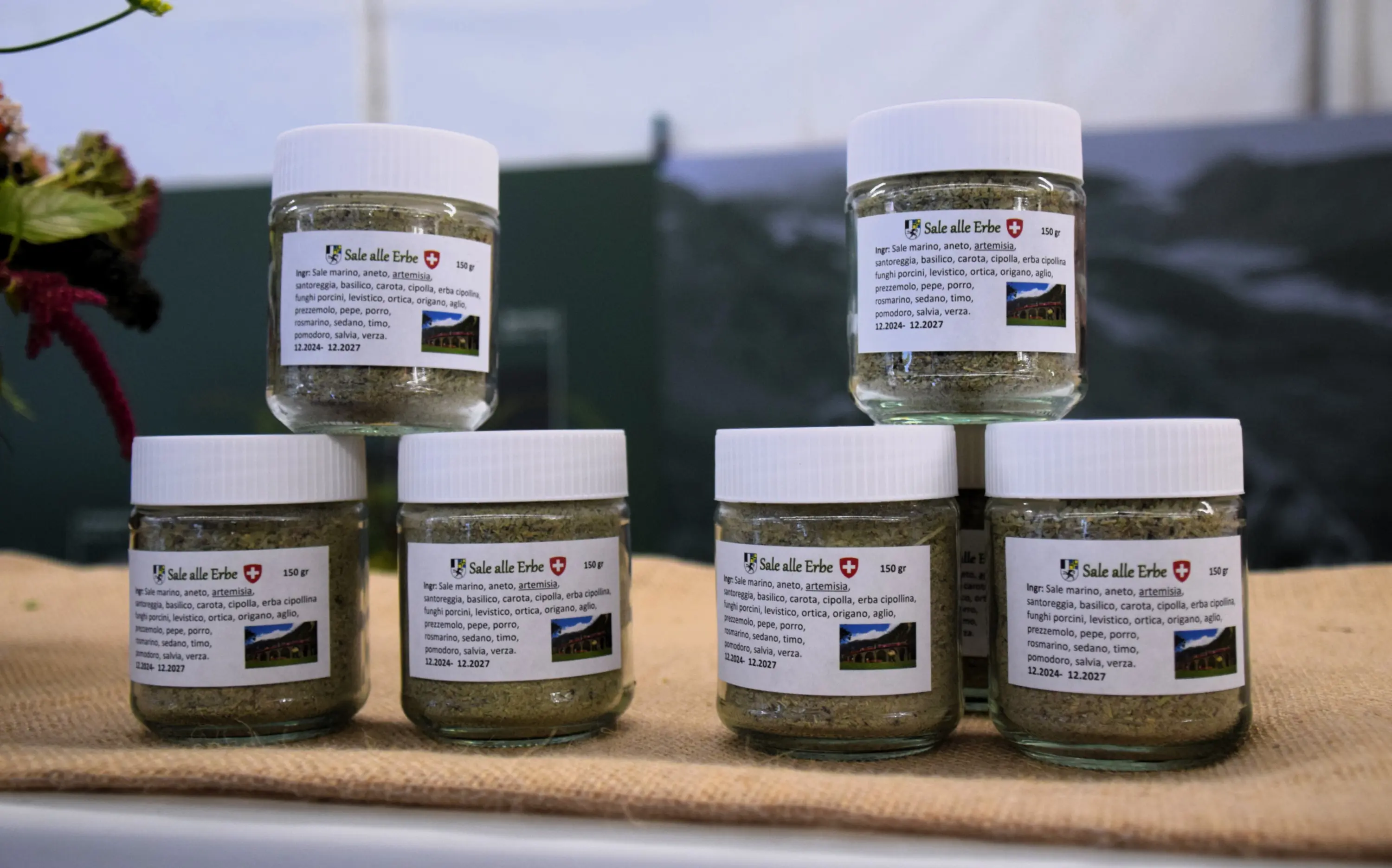 Barattoli di spezie in vetro con etichette, disposti a piramide su un tavolo di juta. I barattoli contengono un mix di erbe aromatiche. Sullo sfondo, una decorazione verde sfocata.