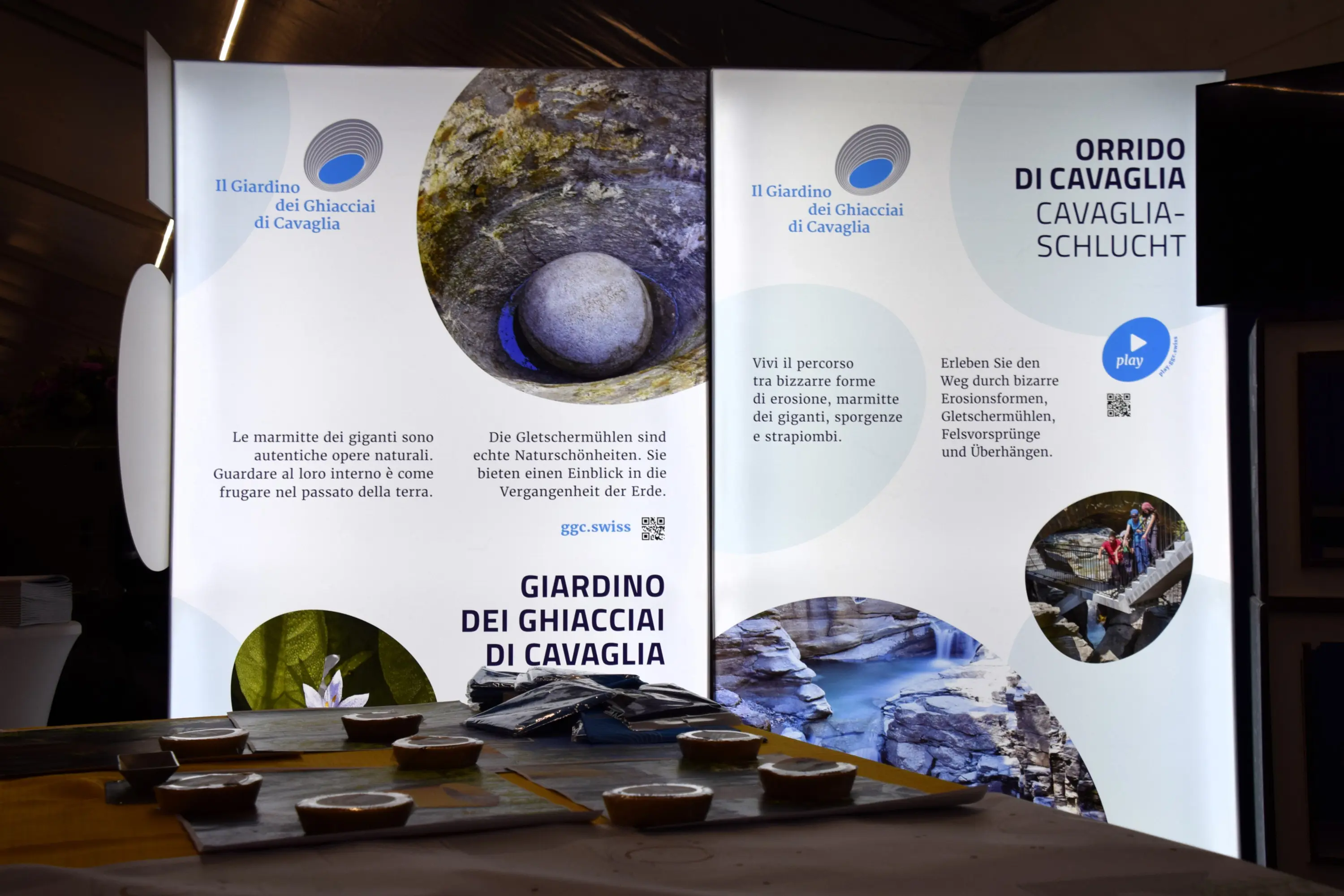 Due pannelli informativi su un'esposizione, con testi in italiano e tedesco riguardanti il Giardino dei Ghiacciai di Cavaglia. Un tavolo davanti mostra vari contenitori in una presentazione.