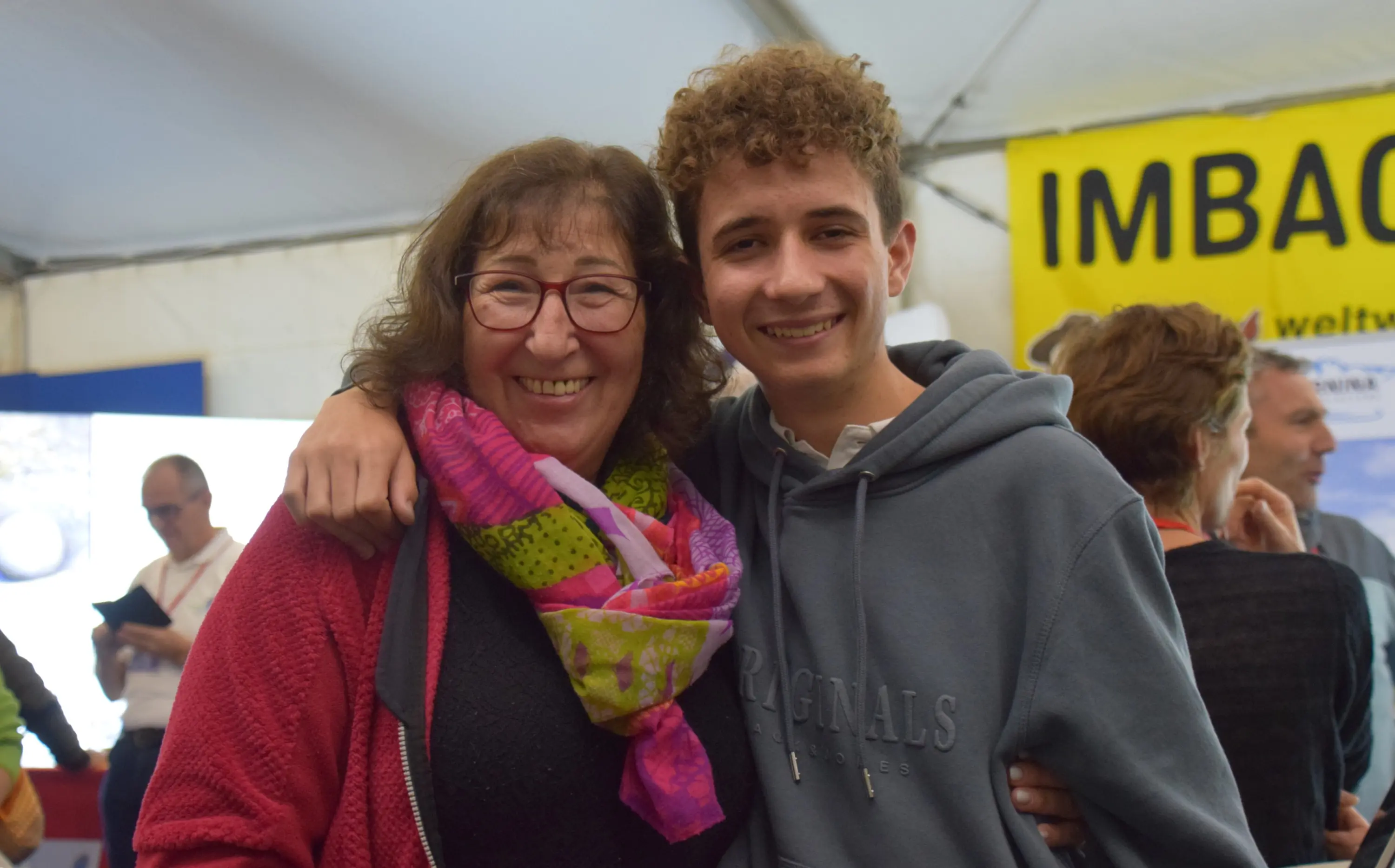 Due persone sorridenti, una donna con occhiali e capelli mossi, e un ragazzo con capelli ricci. Sono abbracciati in un ambiente festoso con poster e gente sullo sfondo.