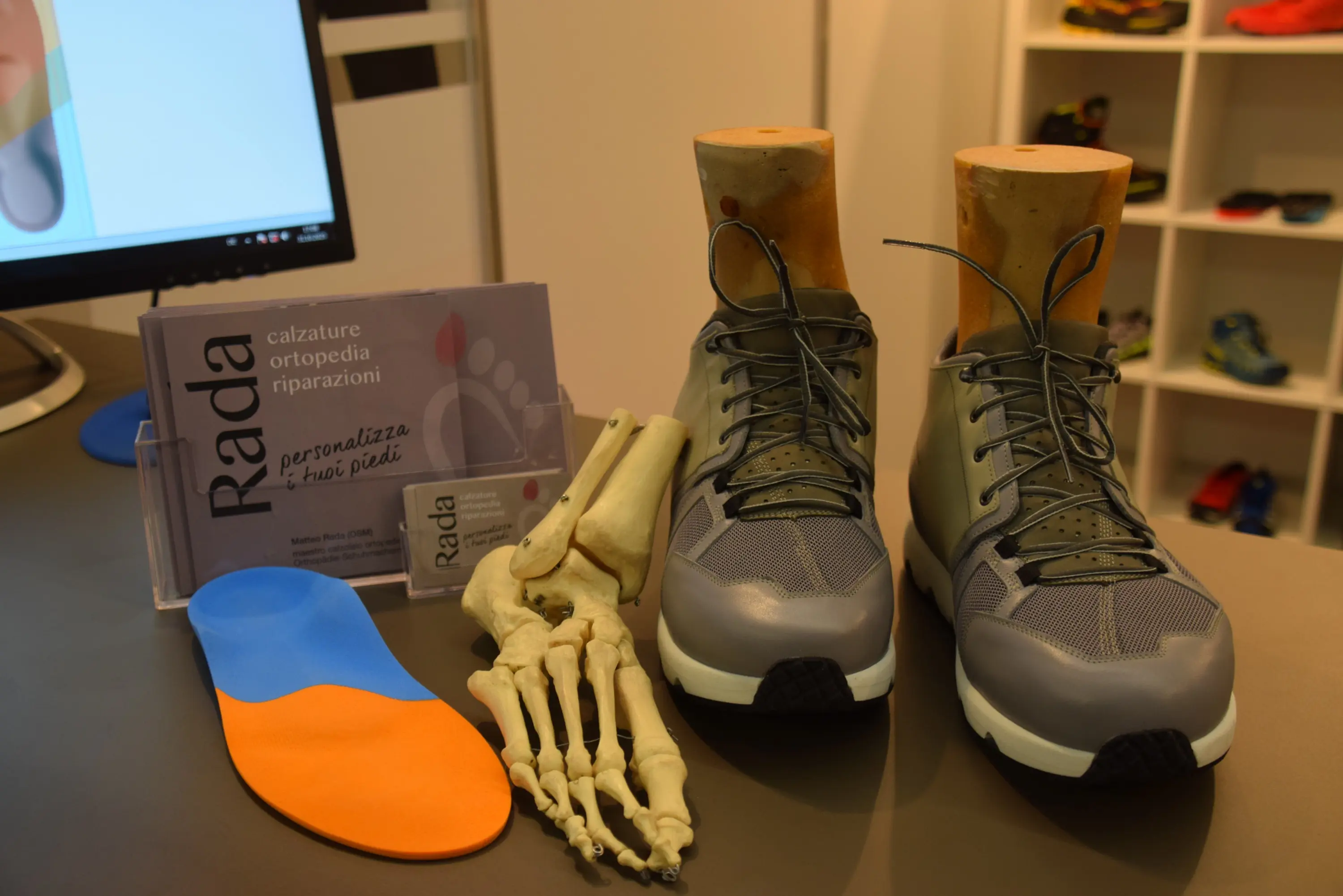 Scarpe da corsa grigie con una forma di legno al posto della soletta, accanto a un modello di scheletro del piede e una soletta arancione e blu. Sullo sfondo si vede un computer e un depliant con il nome "Rada