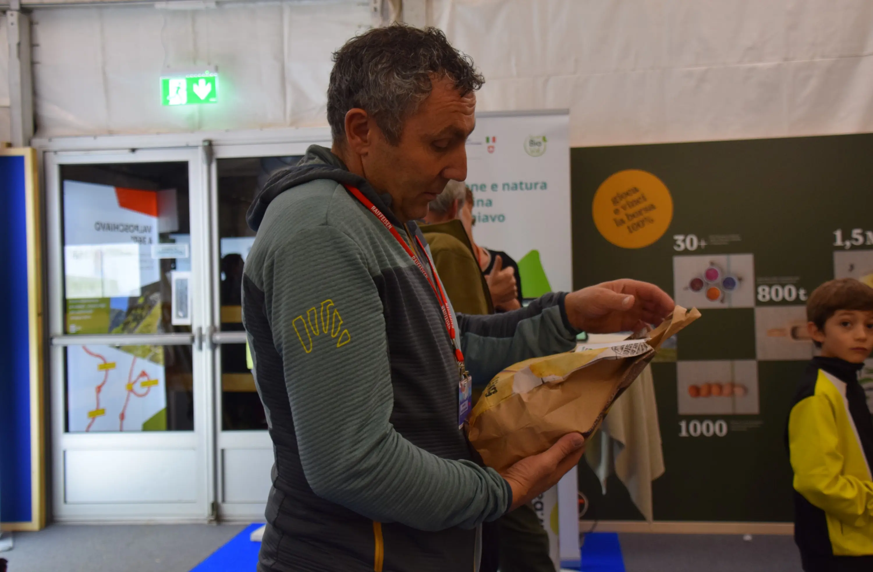Uomo con capelli grigi e ricci, che indossa una giacca verde, mentre esamina un pacchetto all'interno di un tendone espositivo. Un bambino vestito in giallo è vicino a lui sullo sfondo.