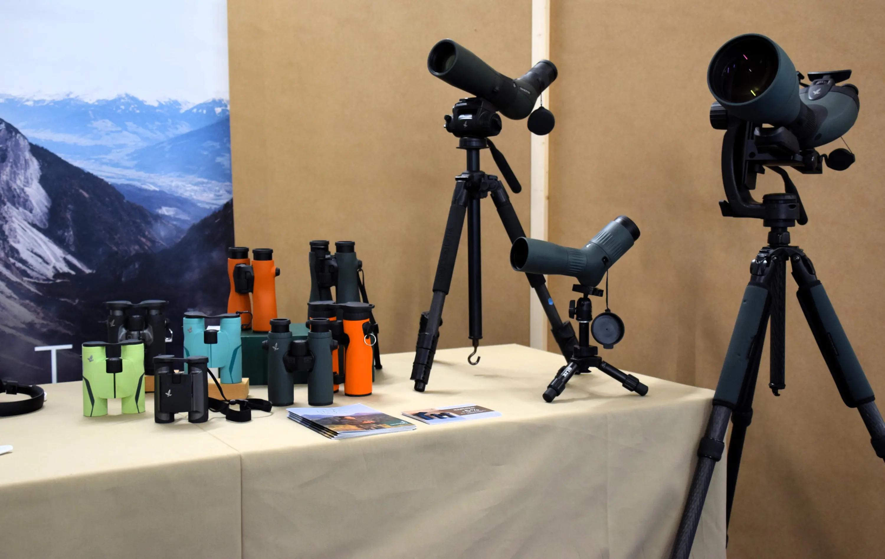 Vari modelli di binocoli e telescopi presentati su un tavolo con uno sfondo montano. Alcuni binocoli sono colorati in verde e arancione, mentre telescopi sono montati su treppiedi. Cartoline informative sono collocate sul tavolo.