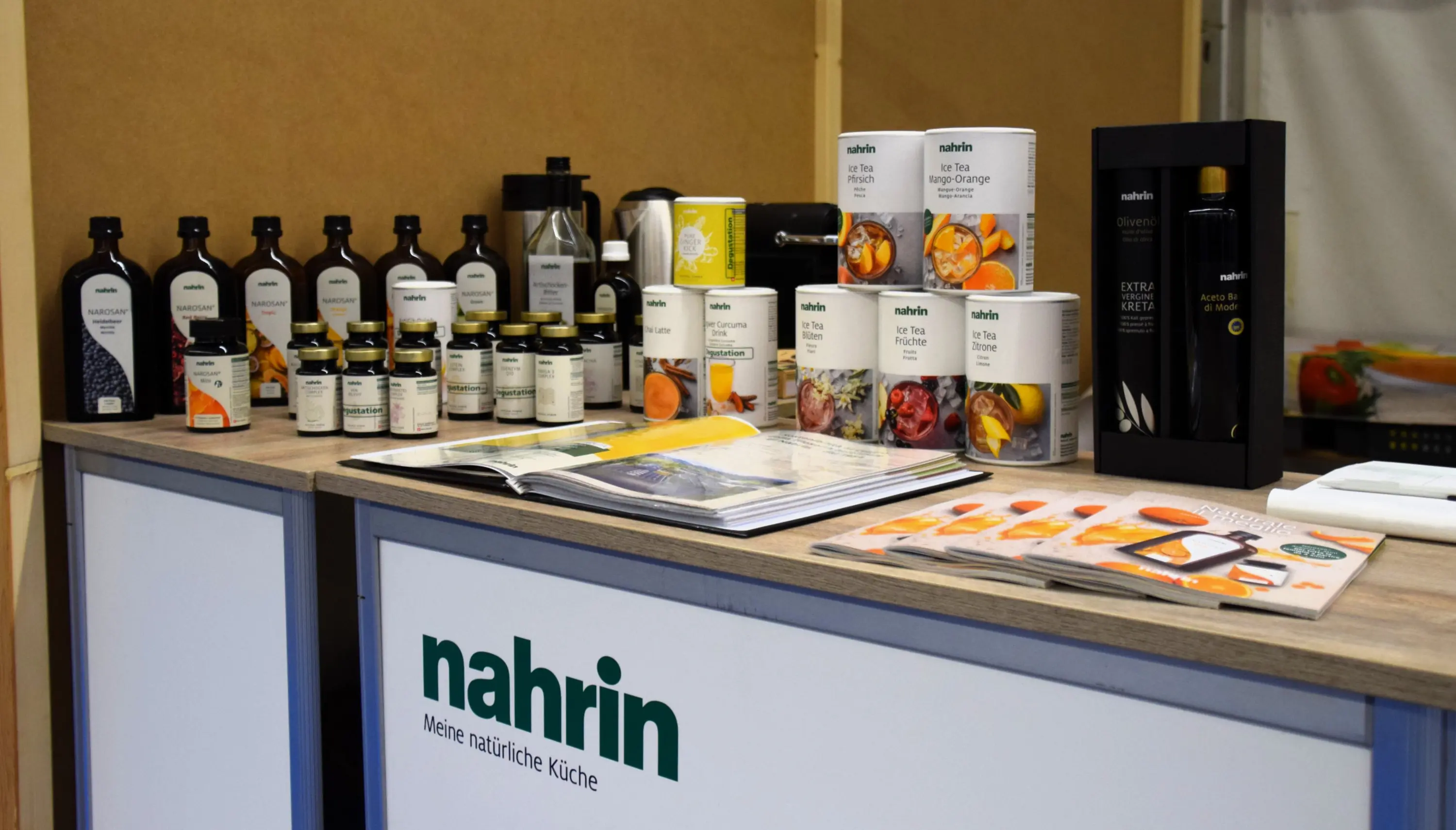 Esposizione di prodotti nahrin su un bancone, inclusi oli, spezie e conserve in contenitori. Un libro di ricette è aperto accanto ai prodotti. Il logo nahrin è ben visibile sulla base del bancone.