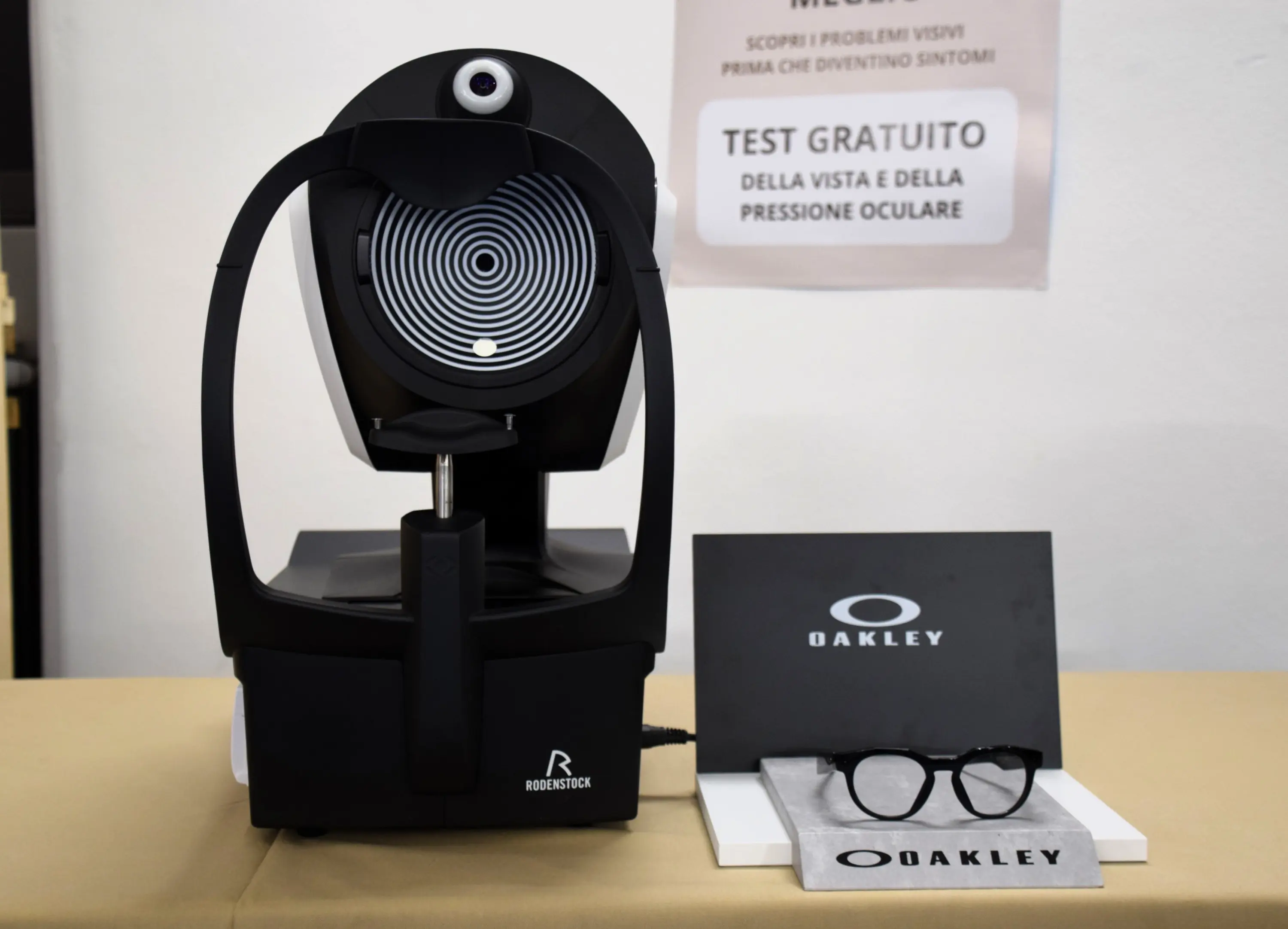 Dispositivo per il test della pressione oculare, posizionato su un tavolo beige, accanto a occhiali Oakley e una brochure informativa.
