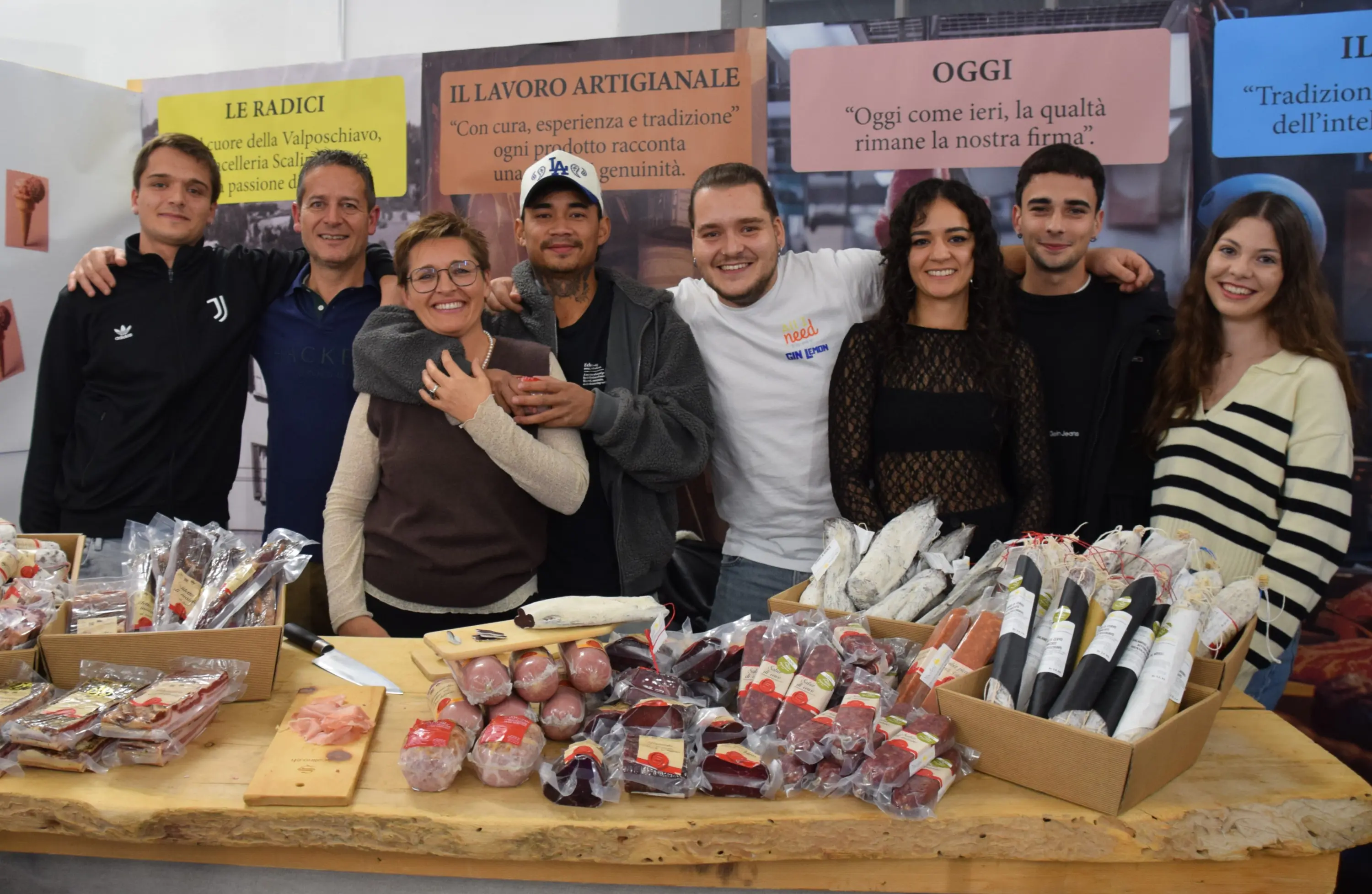 Un gruppo di otto persone sorridenti posa dietro un tavolo carico di salumi e prodotti artigianali. Alcuni membri indossano magliette con il logo di un'azienda. Sullo sfondo, si vedono banner con scritte che evidenziano trad