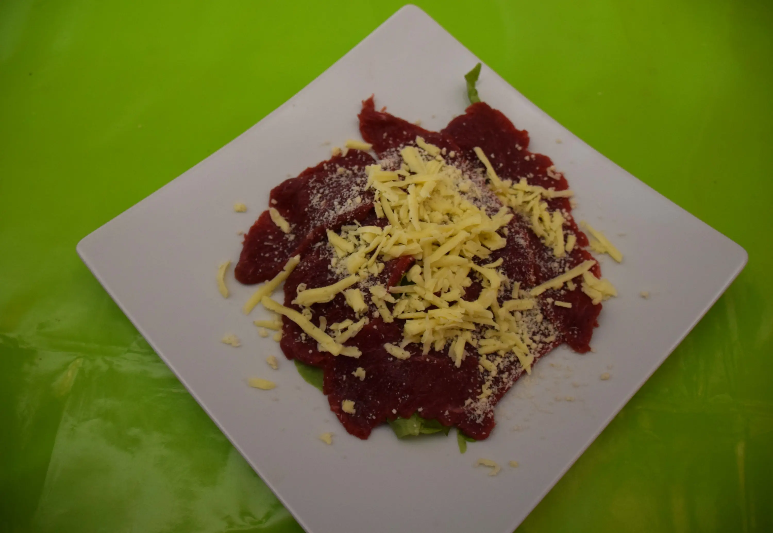 Un piatto di carne cruda, con fettine di carne di manzo sottili, adagiate su un letto di insalata, cosparse di formaggio grattugiato. Il piatto è servito su un fondo verde luminoso.