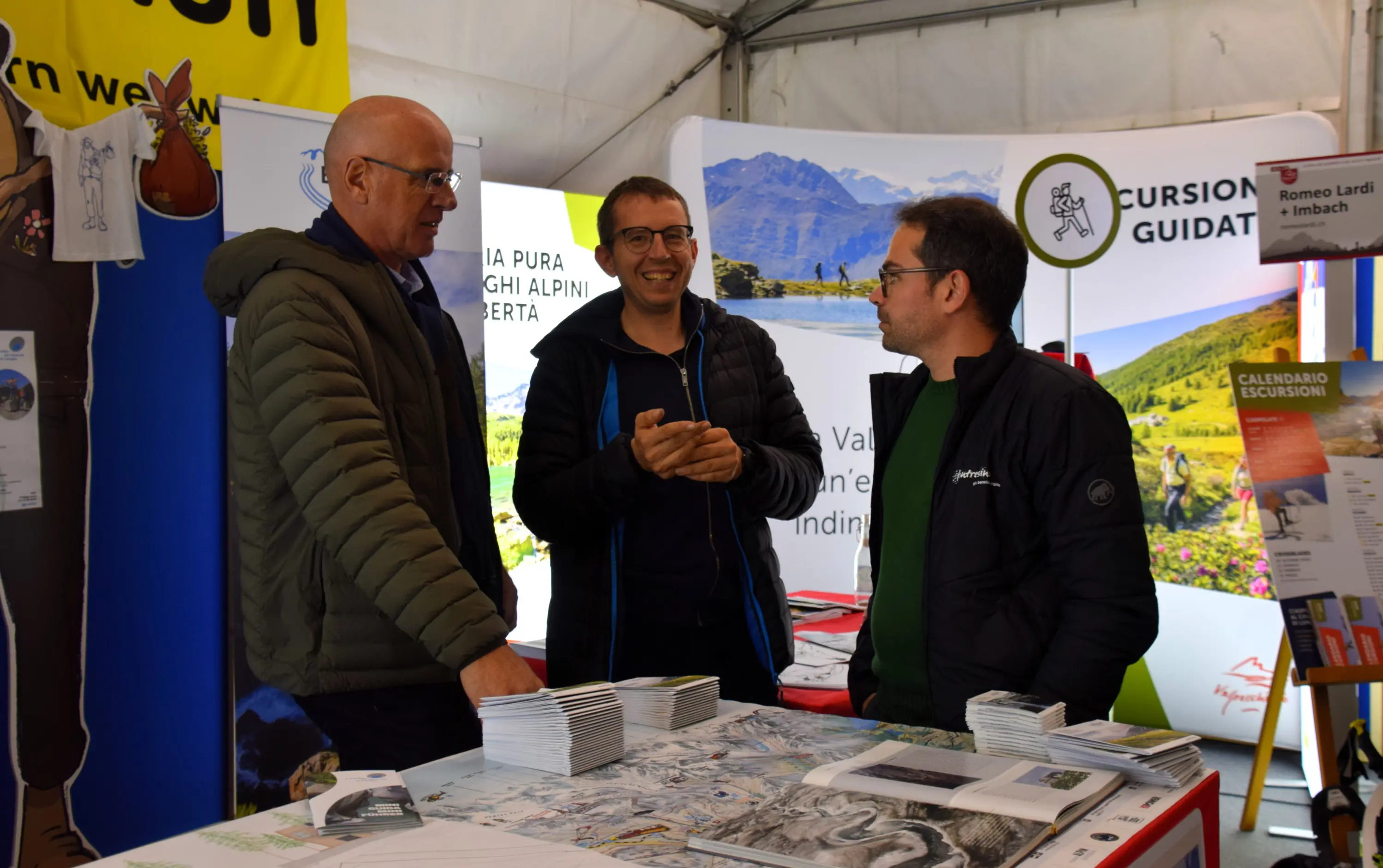 Tre persone sorridenti discutono davanti a un tavolo pieno di materiale informativo su escursioni e itinerari. Sullo sfondo si vedono poster e brochure legate al turismo e alla natura.