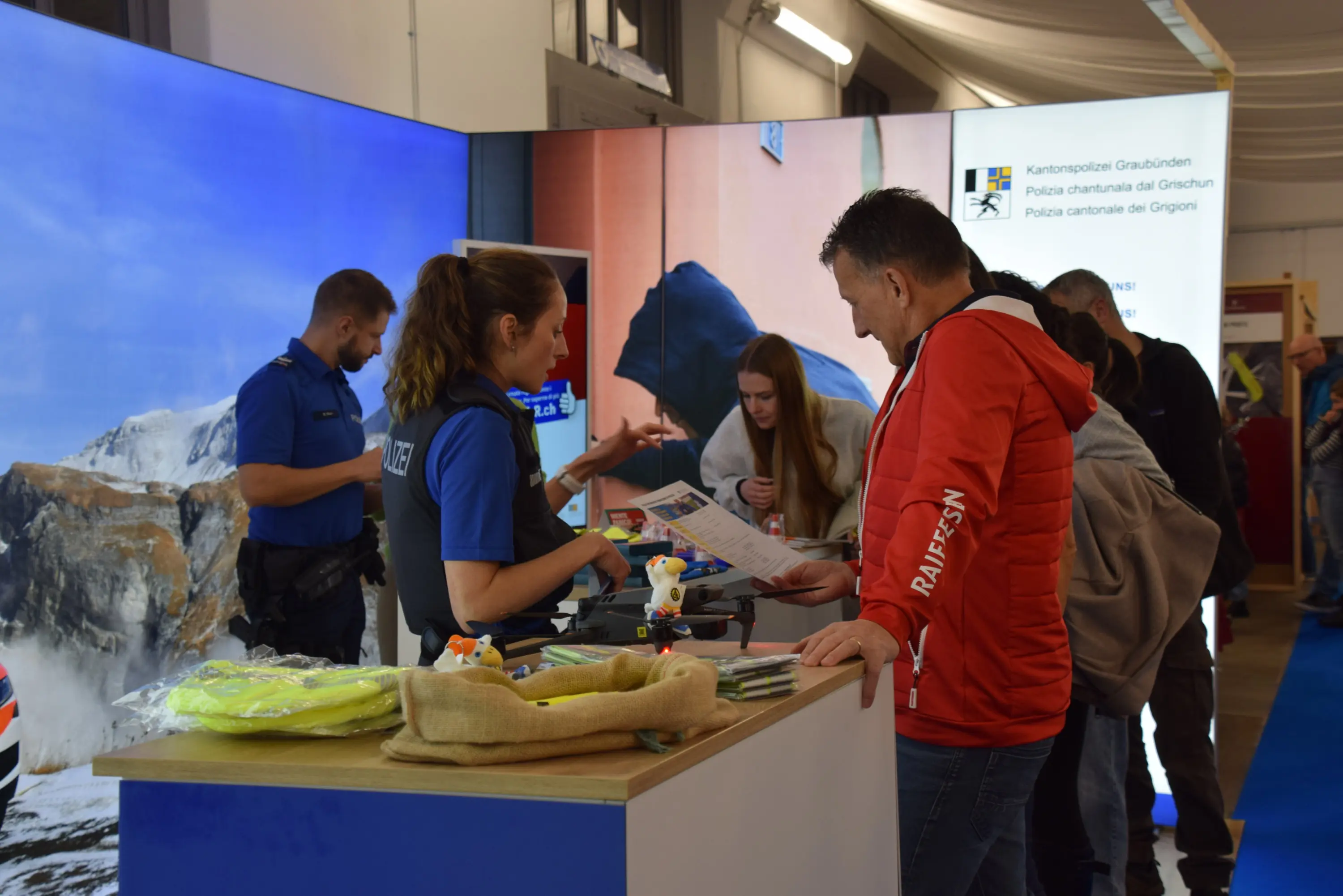 Stand informativo con persone che interagiscono. Una donna in uniforme spiega ai visitatori, mentre un uomo in giacca rossa guarda dei materiali. Sullo sfondo si vedono immagini di montagne.