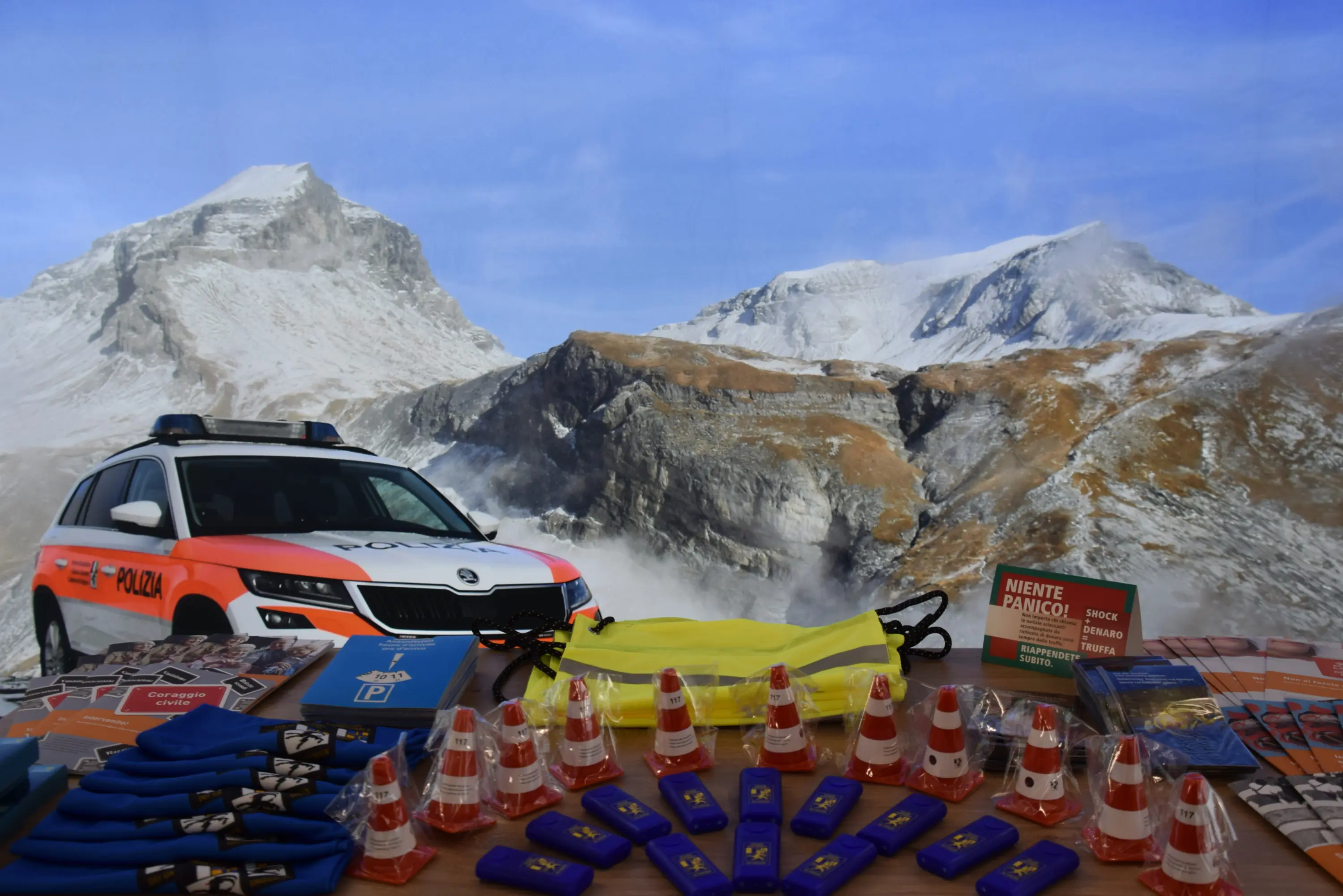 Una macchina della polizia parcheggiata davanti a una montagna innevata. Sul tavolo ci sono vari oggetti, tra cui coni stradali, zaini gialli e brochure informative.