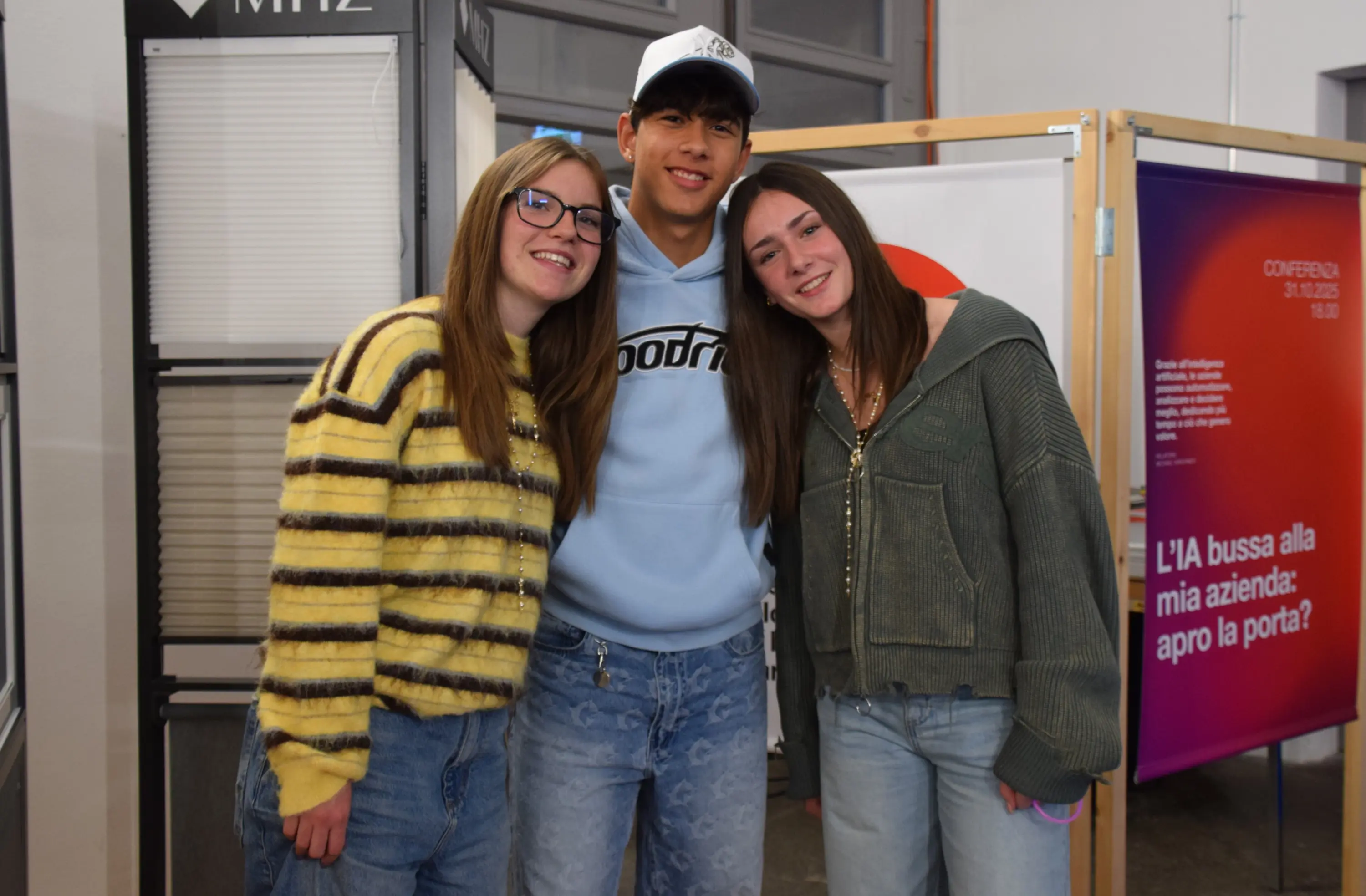 Tre giovani persone sorridono insieme, indossando abbigliamento casual. Una ragazza ha capelli lunghi e castani, l'altra ha capelli biondi. Il ragazzo al centro indossa un cappellino e una felpa. Sullo sfondo si intravede