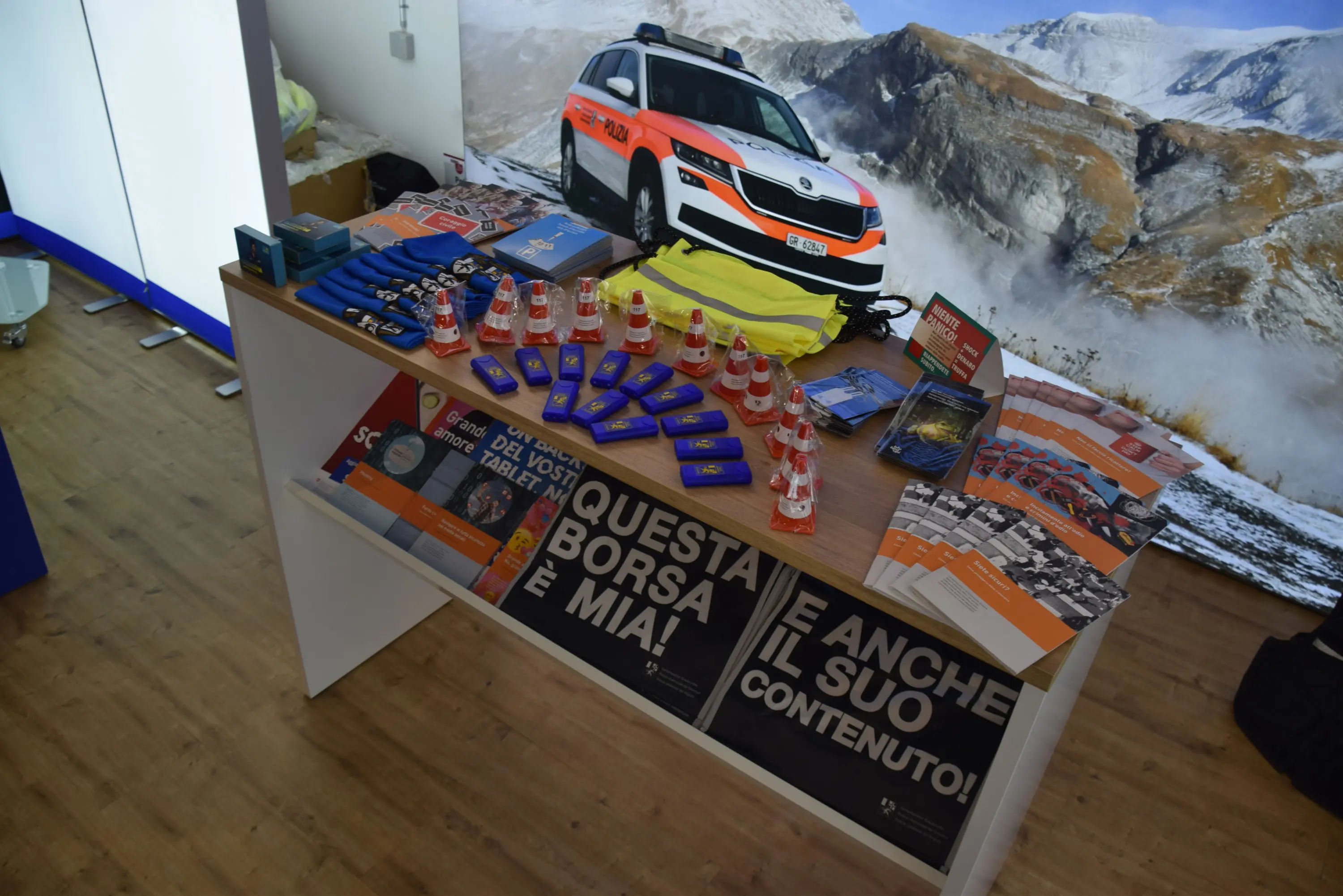 Un tavolo espositivo con vari materiali promozionali, tra cui brochure, penne, caramelle e gadget, con uno sfondo di un'auto di emergenza in montagna.