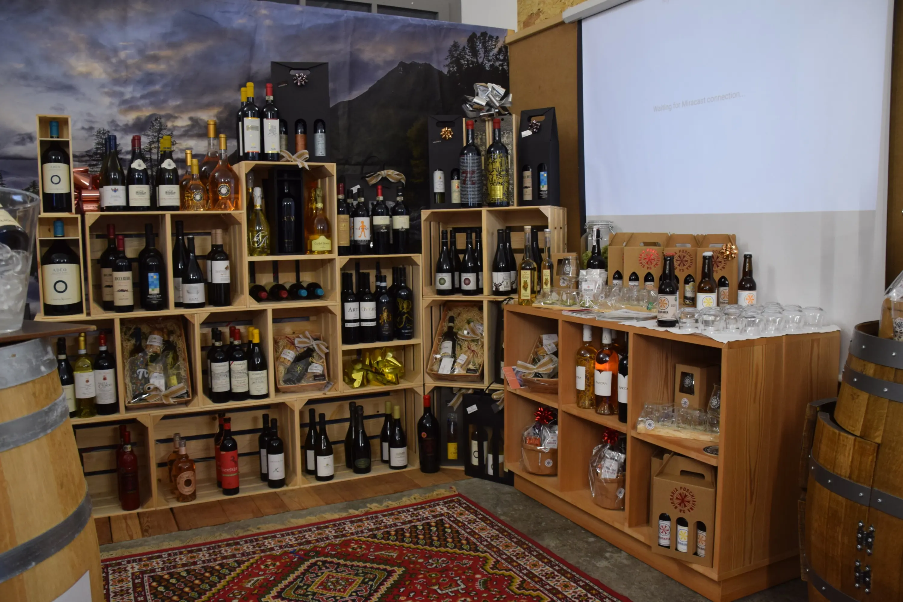 Scaffali in legno contenenti una varietà di bottiglie di vino, champagne e liquori. Un tavolo con bicchieri e un proiettore sullo sfondo. Ambientazione calda e accogliente.