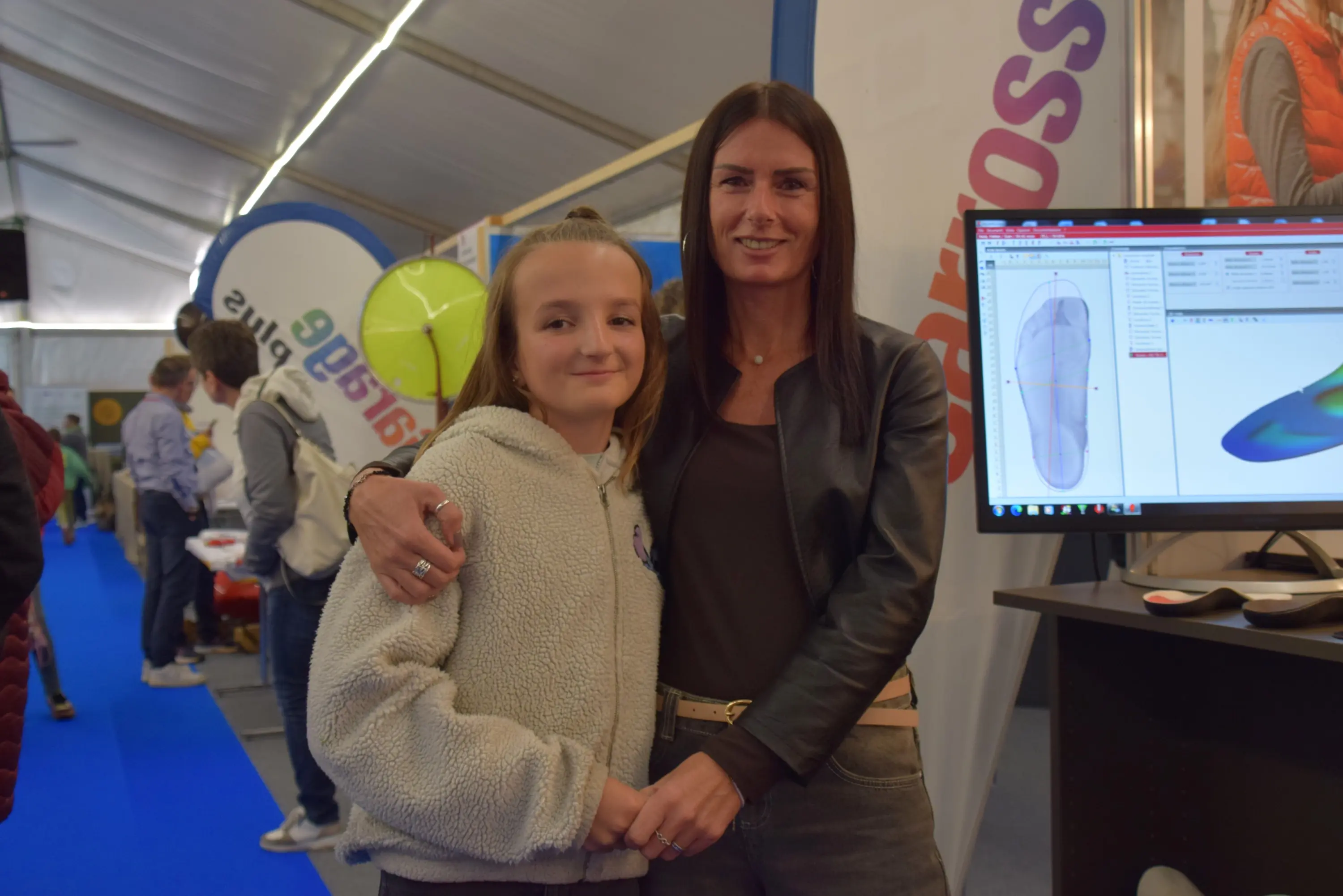 Due donne sorridenti abbracciate in un evento espositivo. Una delle donne è più giovane e indossa un cappotto chiaro, mentre l'altra ha capelli lunghi e scuri e indossa una giacca nera. Sullo sfondo si vedono schermi e altri visit