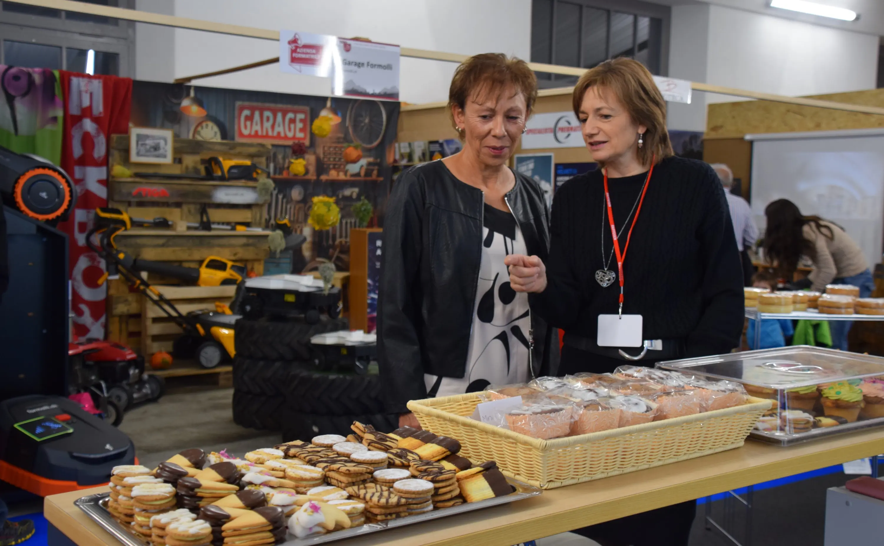 Due donne osservano un vassoio di dolci in una fiera, con un ambiente colorato e diverse attrezzature visibili sullo sfondo.