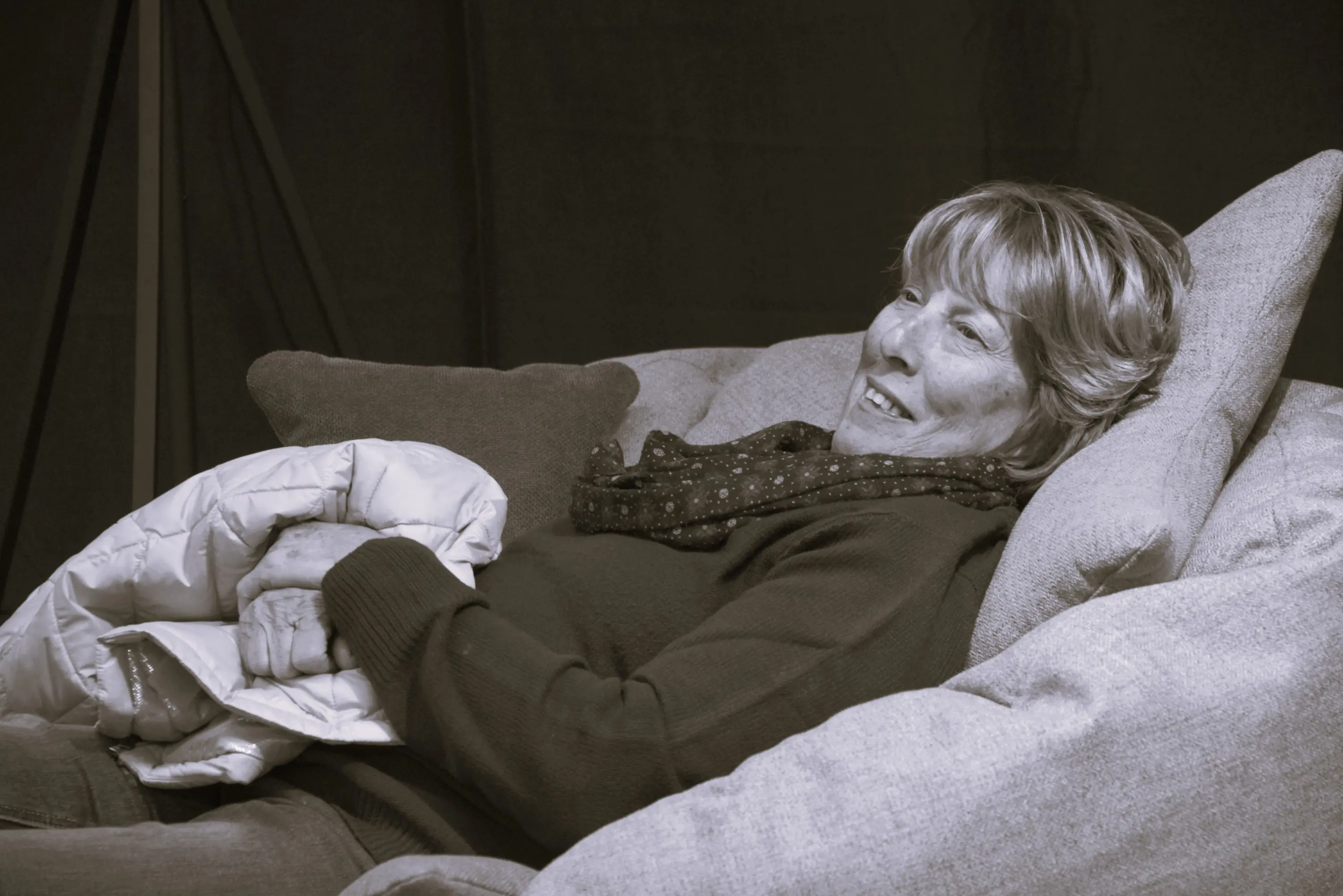 Una donna sdraiata su un divano, sorridente, con una coperta nelle mani e uno scialle. Lo sfondo è scuro e il contesto sembra rilassante.