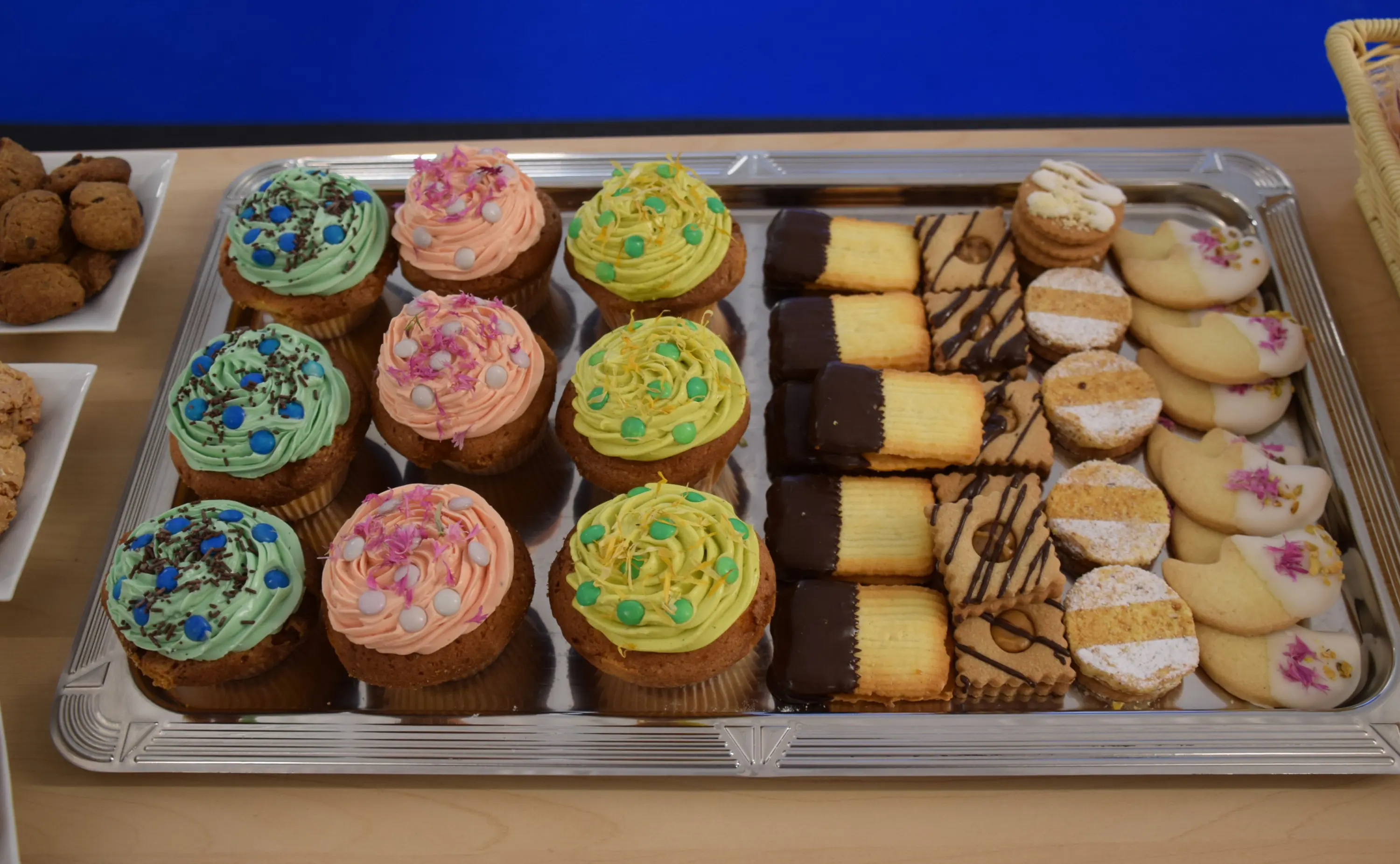 Vassoi di dolci con muffin decorati in azzurro, rosa e giallo, affiancati da biscotti assortiti, inclusi biscotti con cioccolato e decorazioni floreali.