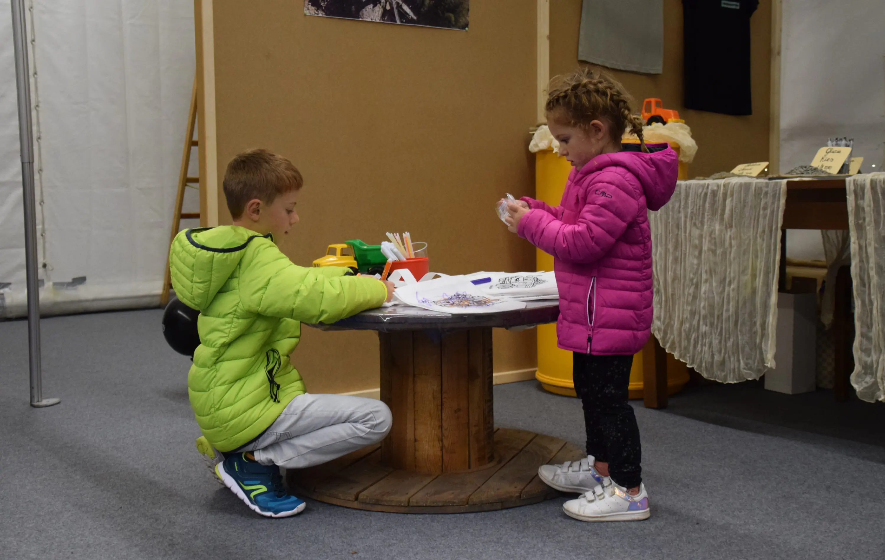 Due bambini, uno con un giubbotto verde e l'altro con un giubbotto rosa, stanno giocando attorno a un tavolo di legno. Sul tavolo ci sono materiali per colorare e giochi.