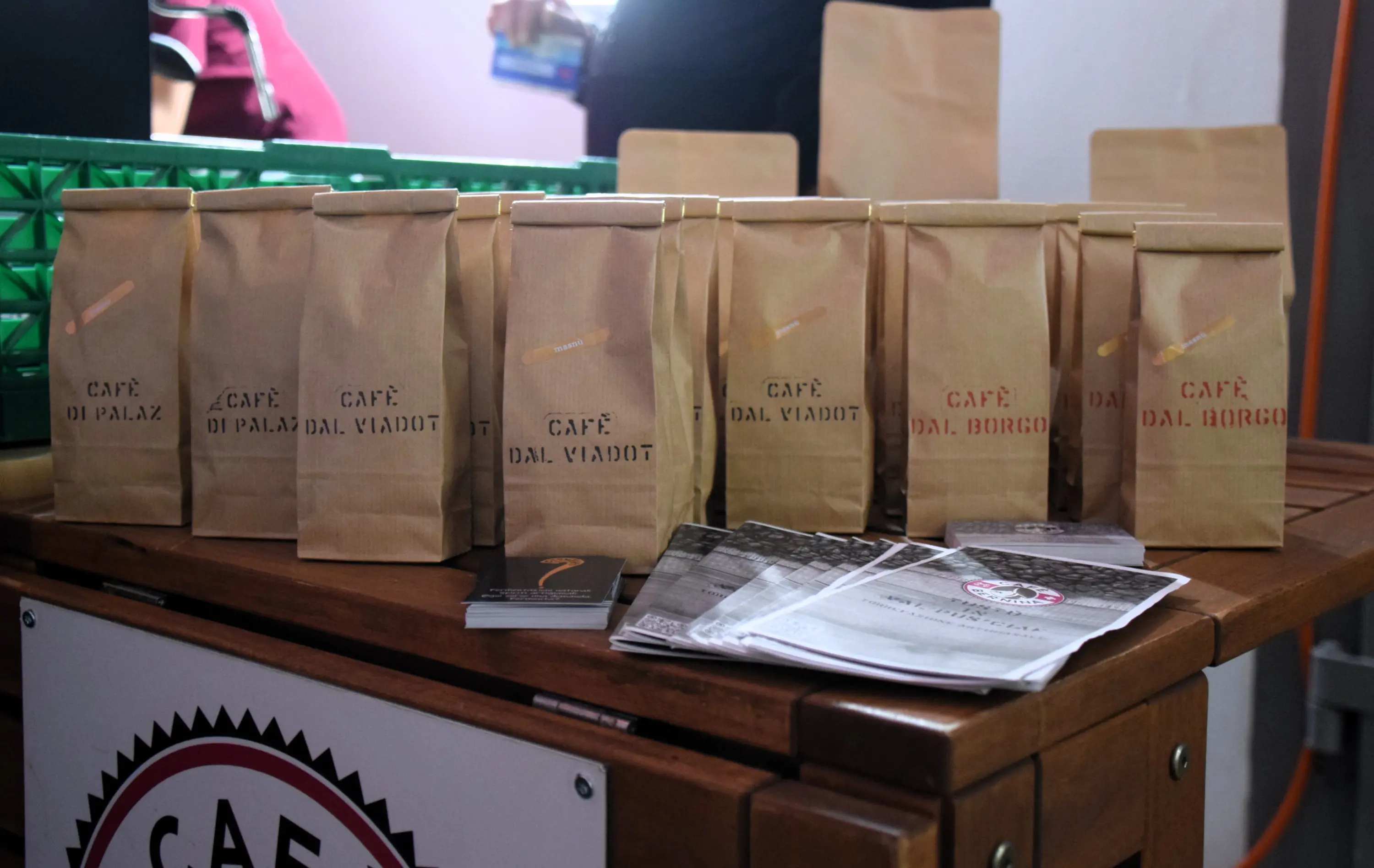 Varie confezioni di caffè in sacchetti di carta, etichettati con nomi come "Café dal Palazzo" e "Café dal Viadotto", disposti su un tavolo di legno. In background, ci sono alcuni volantini.