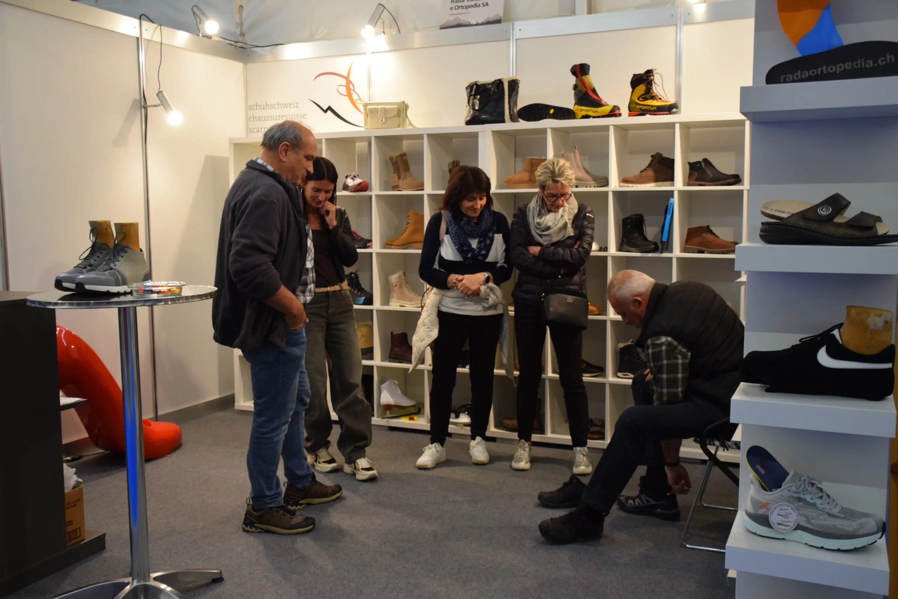 Un gruppo di cinque persone osserva e prova delle scarpe all'interno di uno stand espositivo, circondati da scaffali che mostrano vari modelli di calzature.