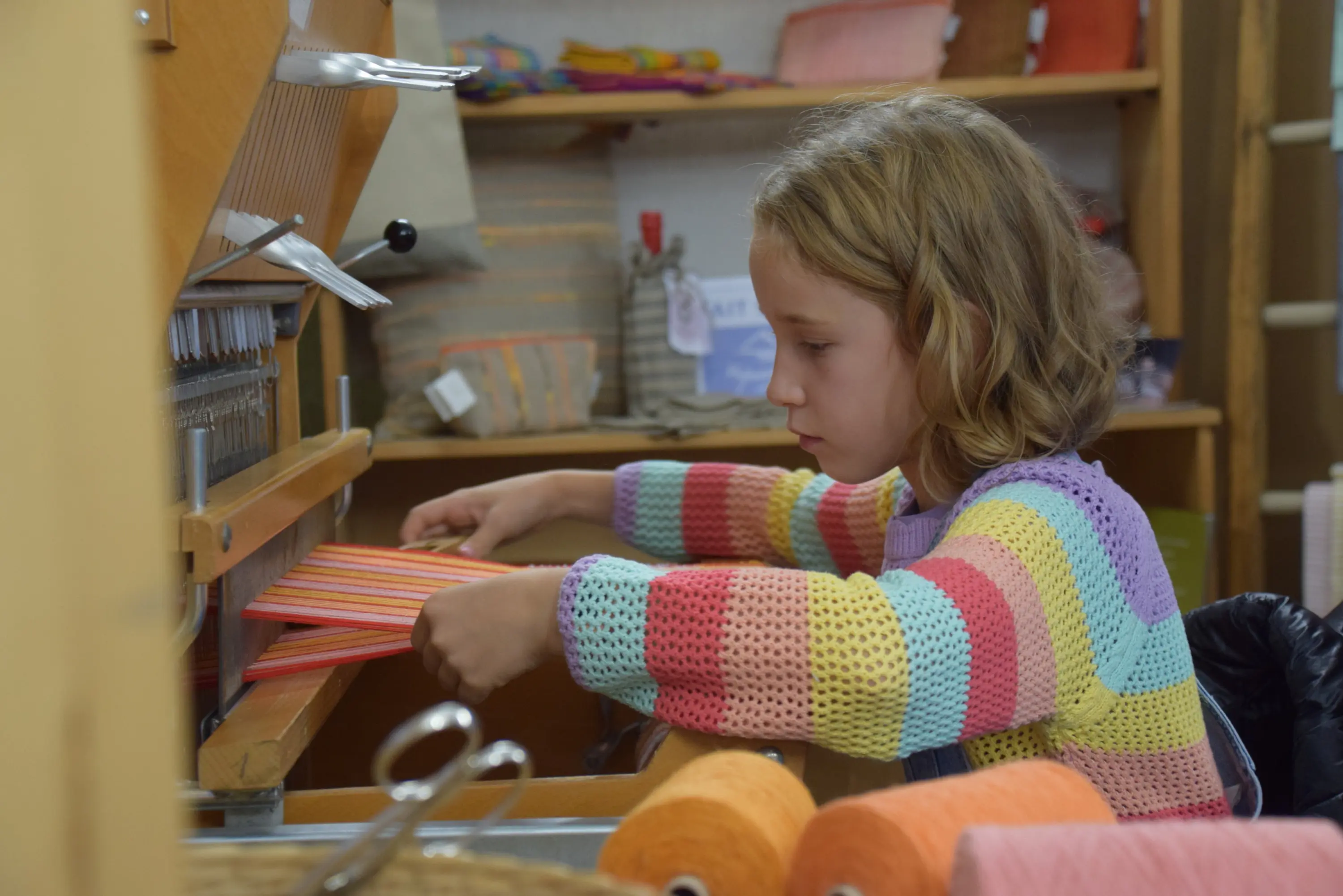Una bambina con un maglione colorato a strisce sta lavorando a un telaio, utilizzando fili di diversi colori. Sullo sfondo si vedono rotoli di filato e oggetti di artigianato.