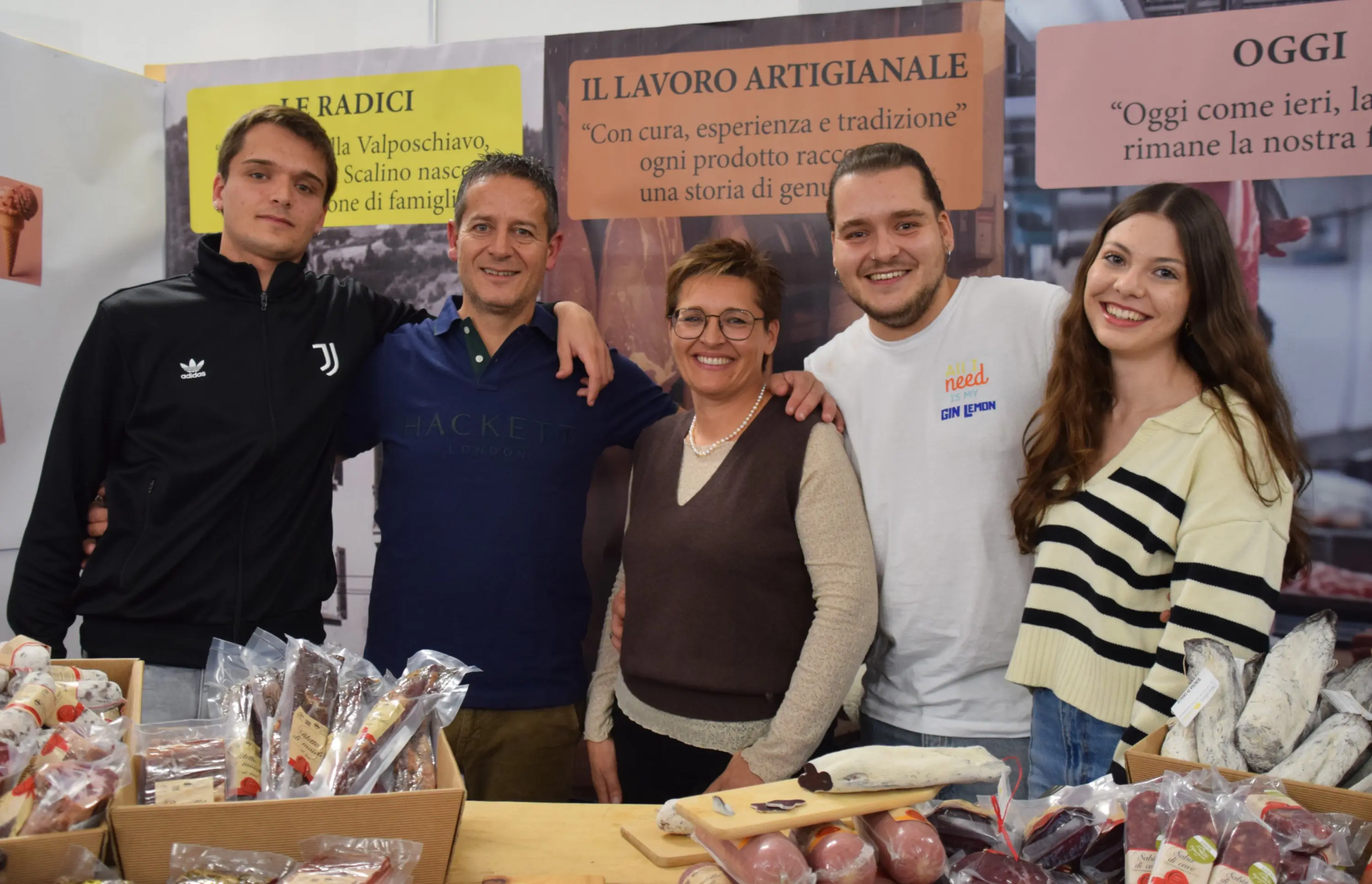 Un gruppo di cinque persone sorridenti si trova davanti a un banco di prodotti artigianali. Sullo sfondo ci sono pannelli informativi con testi in italiano. Il gruppo è composto da due uomini, una donna e due giovani, tutti vestiti in modo casual. I prodotti in espos