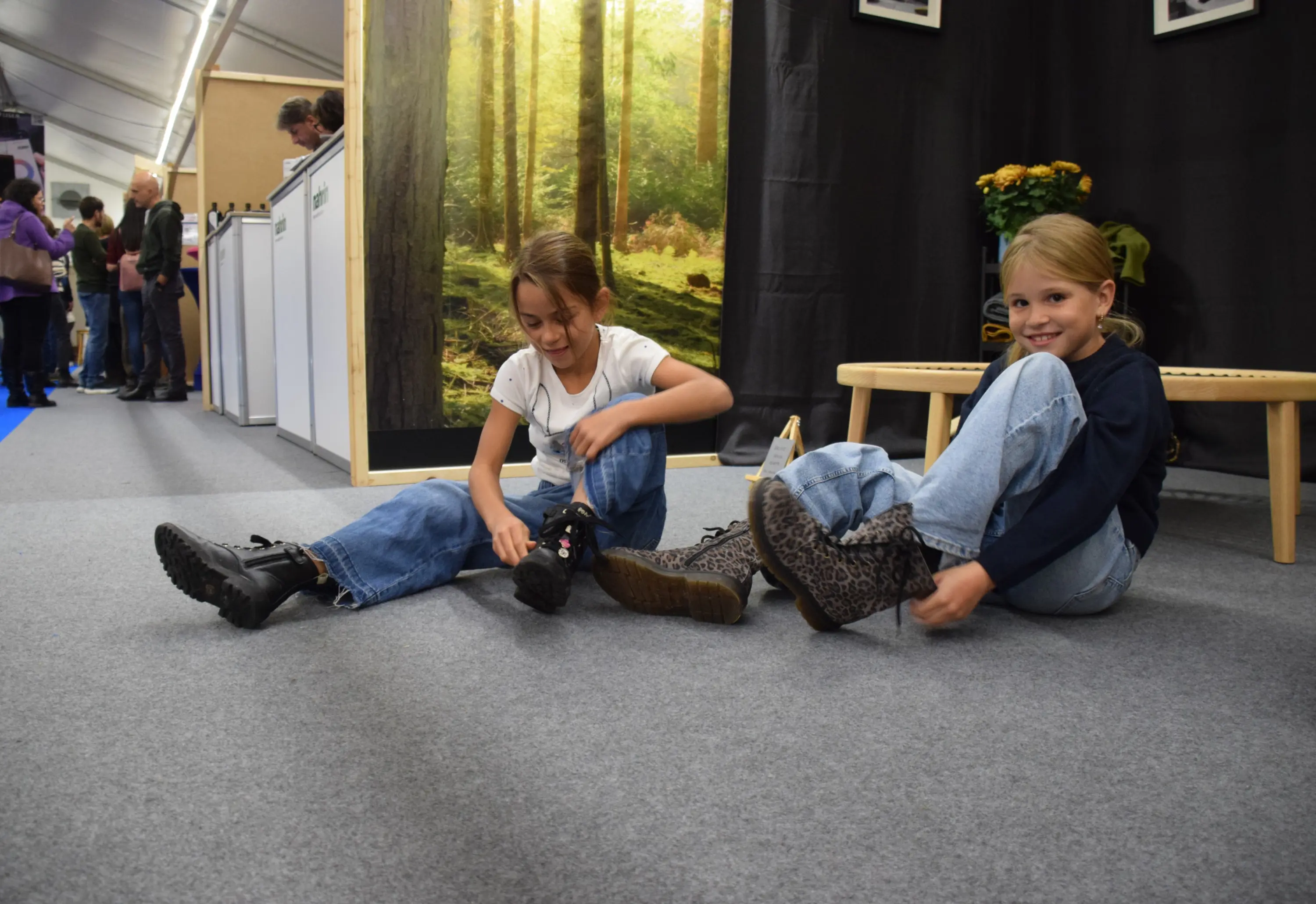Due bambine sorridenti si mettono le scarpe in una fiera. Una indossa stivali neri, l'altra stivali leopardati. Sullo sfondo ci sono stand e persone.