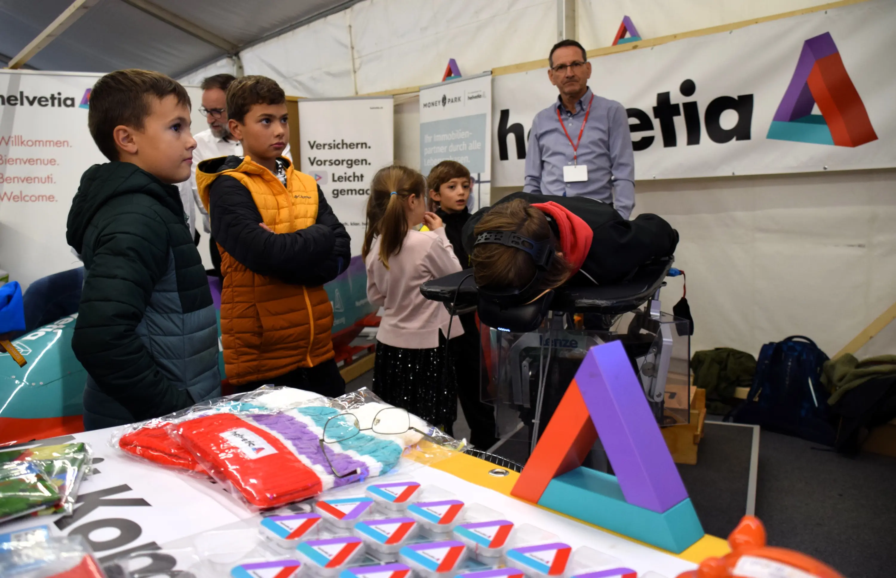 Un gruppo di bambini osserva un dimostratore che utilizza un dispositivo innovativo all'interno di una tenda espositiva. Sul tavolo sono visibili vari oggetti promozionali e materiali informativi.