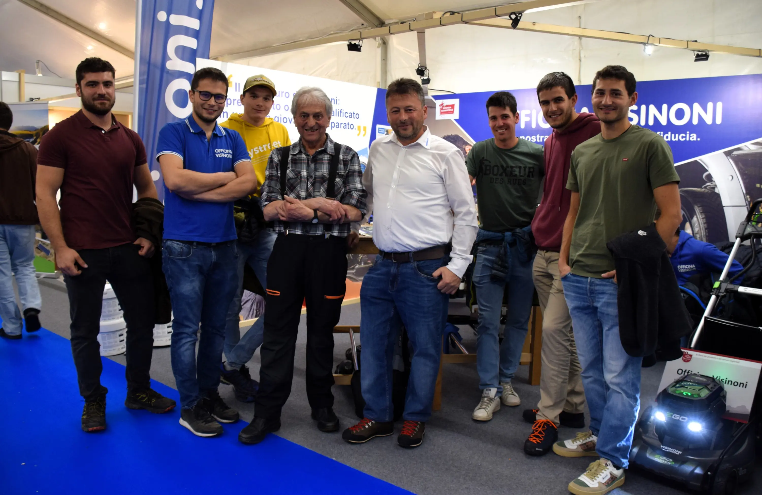 Un gruppo di nove persone sorride e posa insieme in un ambiente fieristico. Alcuni indossano magliette colorate e jeans, mentre uno è in camicia e pantaloni formali. Sullo sfondo si vedono banner con nomi e informazioni relative all'evento.