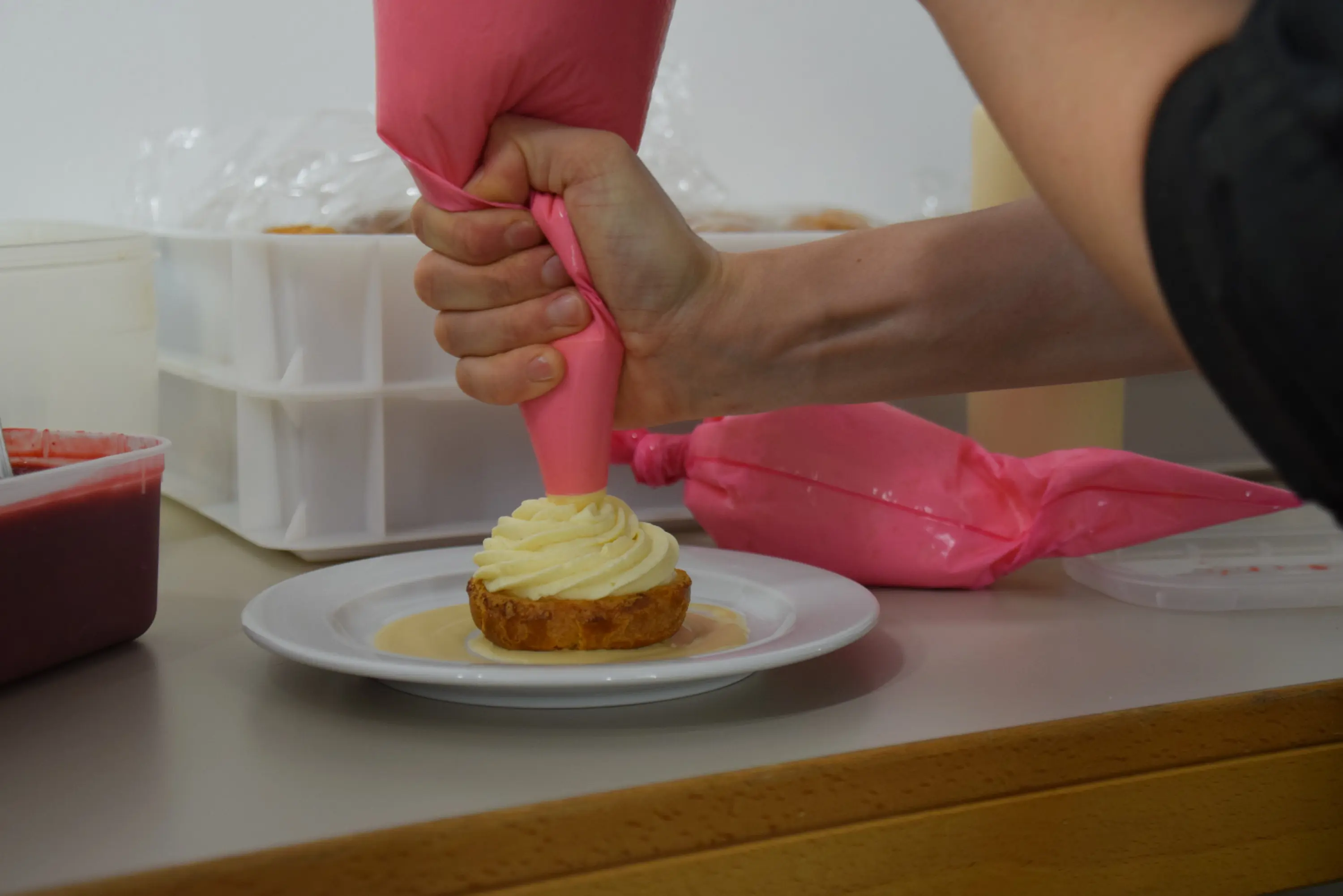 Una mano che utilizza una sac à poche rosa per decorare un dolce con crema su un piatto bianco. Sullo sfondo, ci sono contenitori di ingredienti e sacchetti di crema.