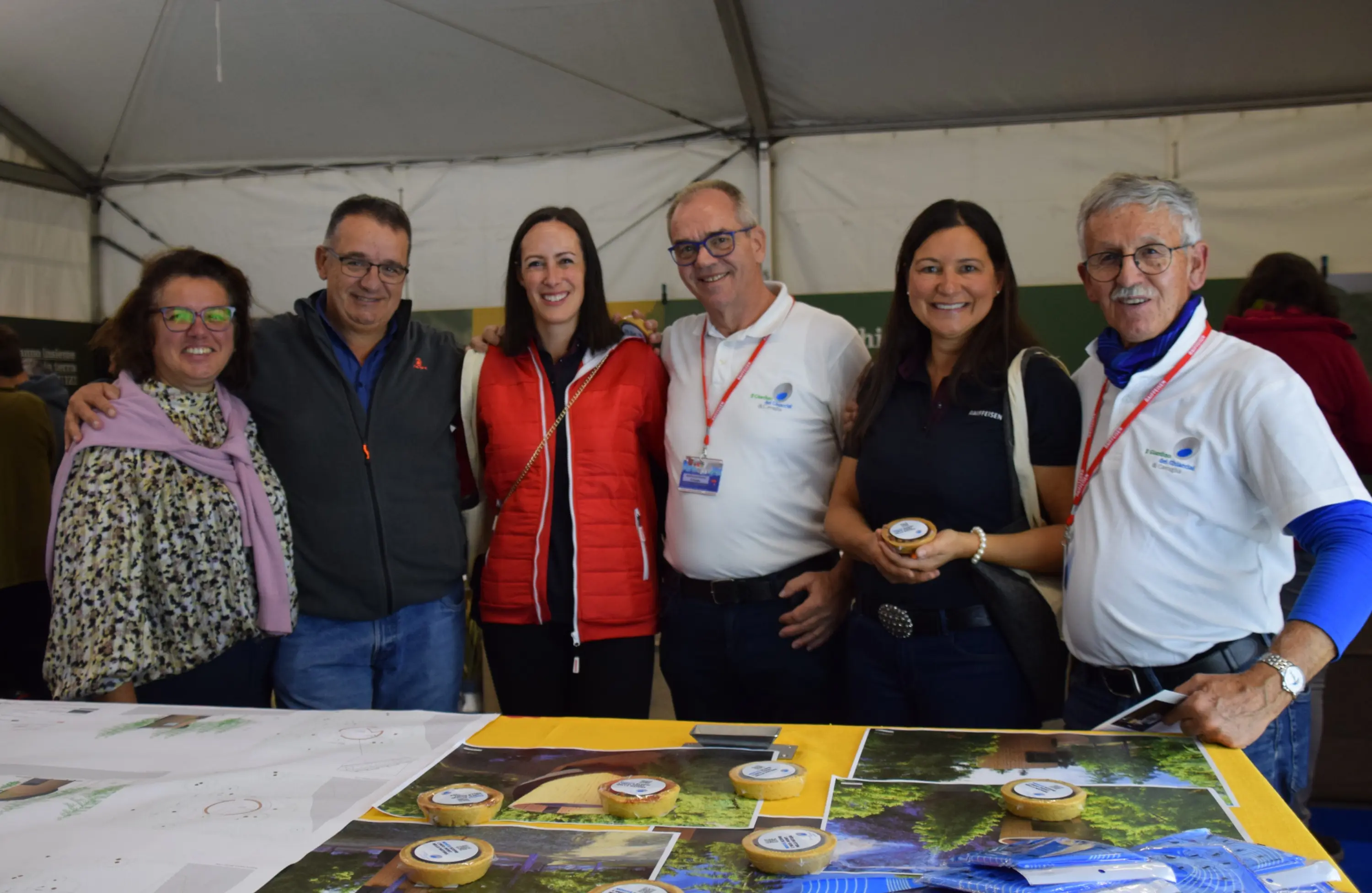 Gruppo di sei persone sorridenti, in un ambiente interno coperto da una tenda. Sono davanti a un tavolo con fotografie e contenitori. Ognuno indossa abiti casual e alcuni hanno distintivi o magliette con un logo.