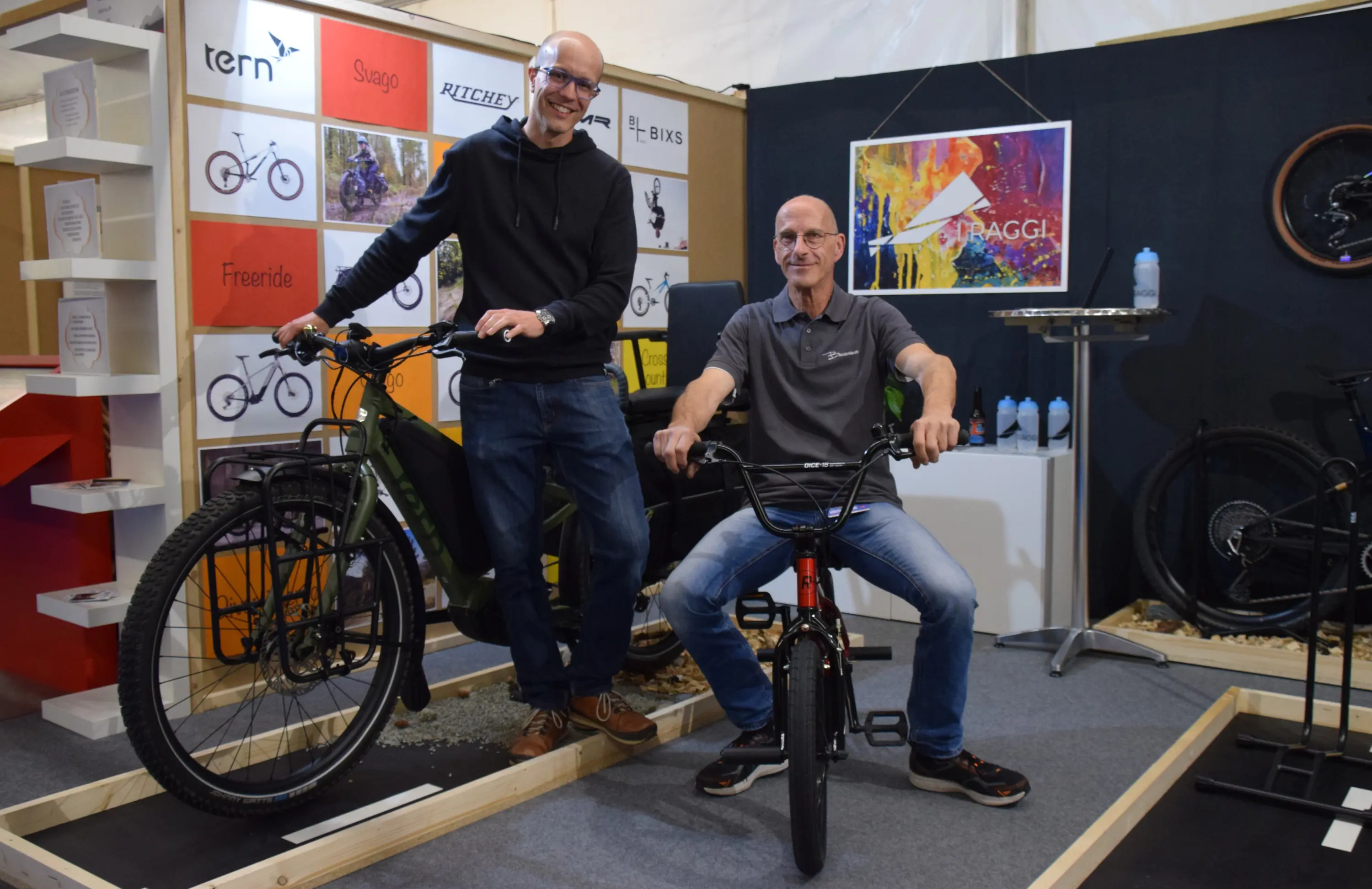 Due uomini sorridenti posano in un'esposizione di biciclette. Uno è in piedi accanto a una mountain bike, mentre l'altro è seduto su una bici gigante BMX. Sullo sfondo si vedono vari modelli di biciclette esposti.
