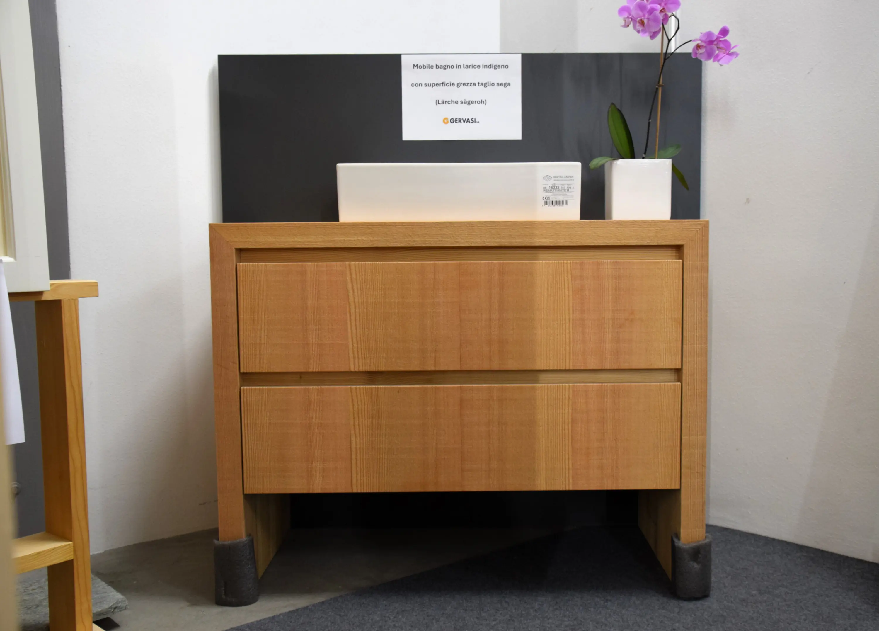 Cassettiera in legno chiaro con due cassetti, posizionata accanto a un pannello scuro. Su di esso c'è un vaso con un'orchidea e un cartello informativo.