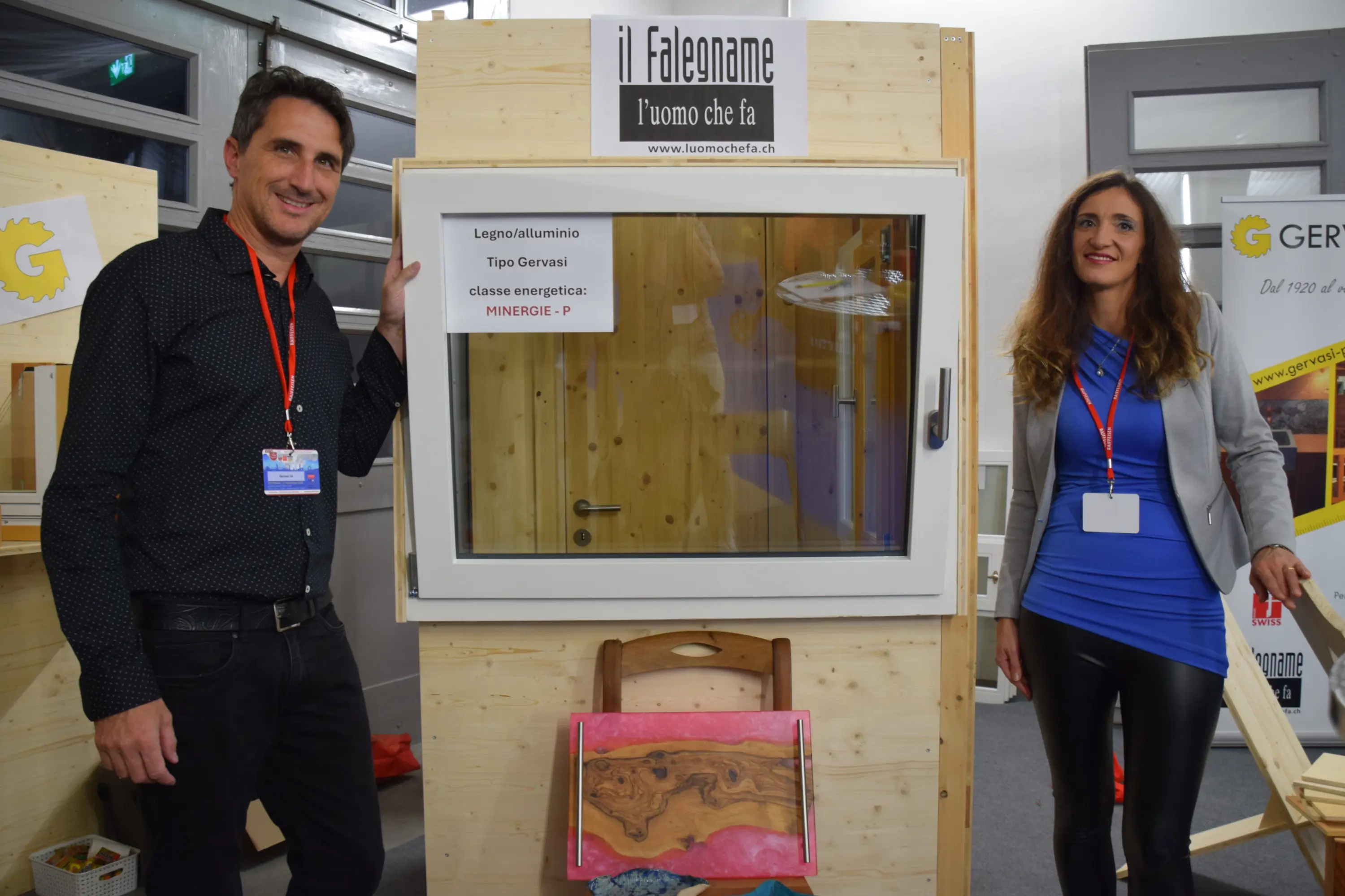 Due persone sorridenti, un uomo e una donna, stanno posizionati accanto a una finestra moderna in legno e alluminio. Sullo sfondo si vedono pannelli di legno e un cartello che recita "Il Falegname - l'uomo che