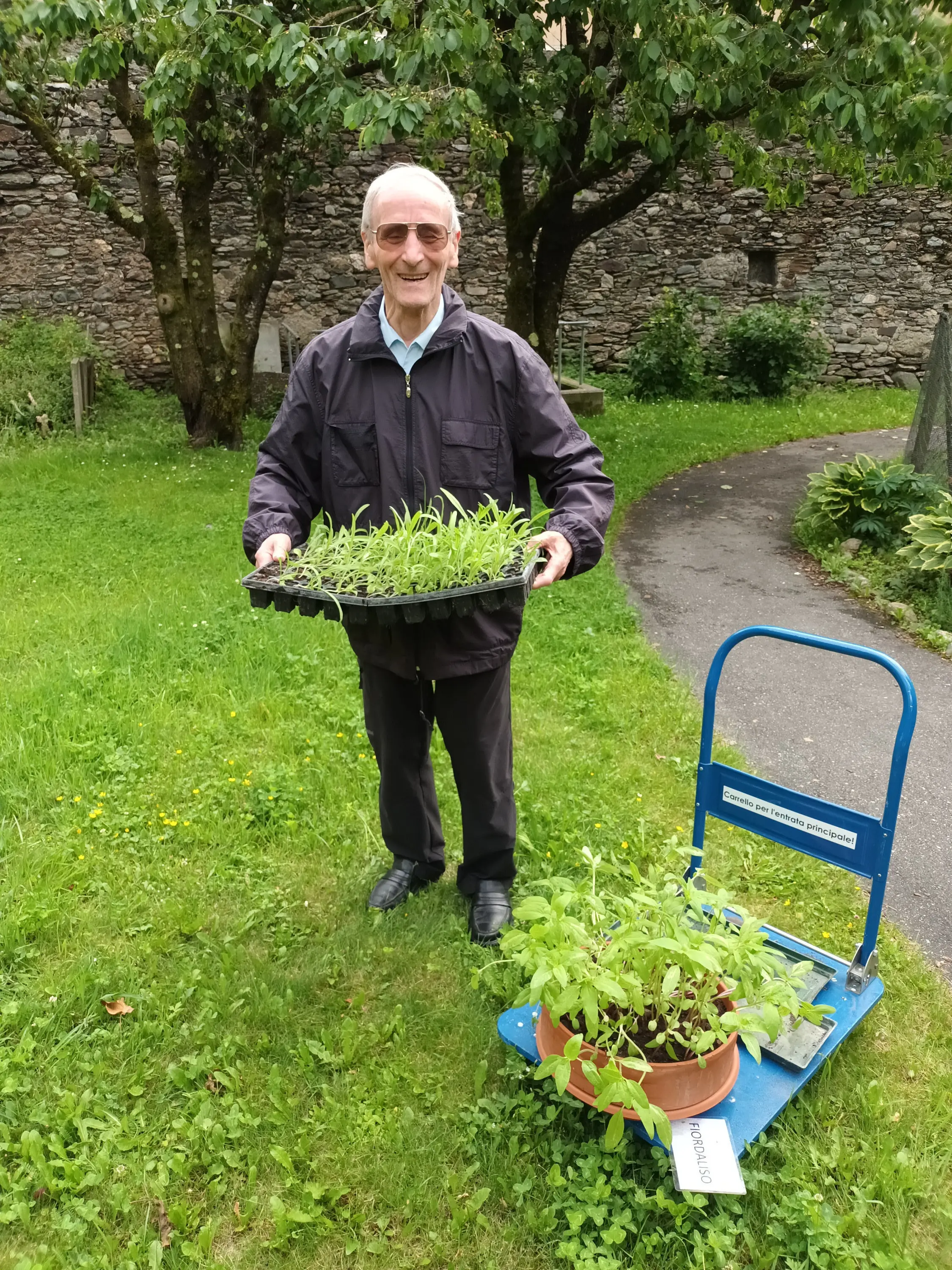 Un uomo sorridente tiene in mano un vassoio di piantine verdi, con un carrello blu carico di piante accanto a lui. Sul terreno erboso, si vedono alberi sullo sfondo.