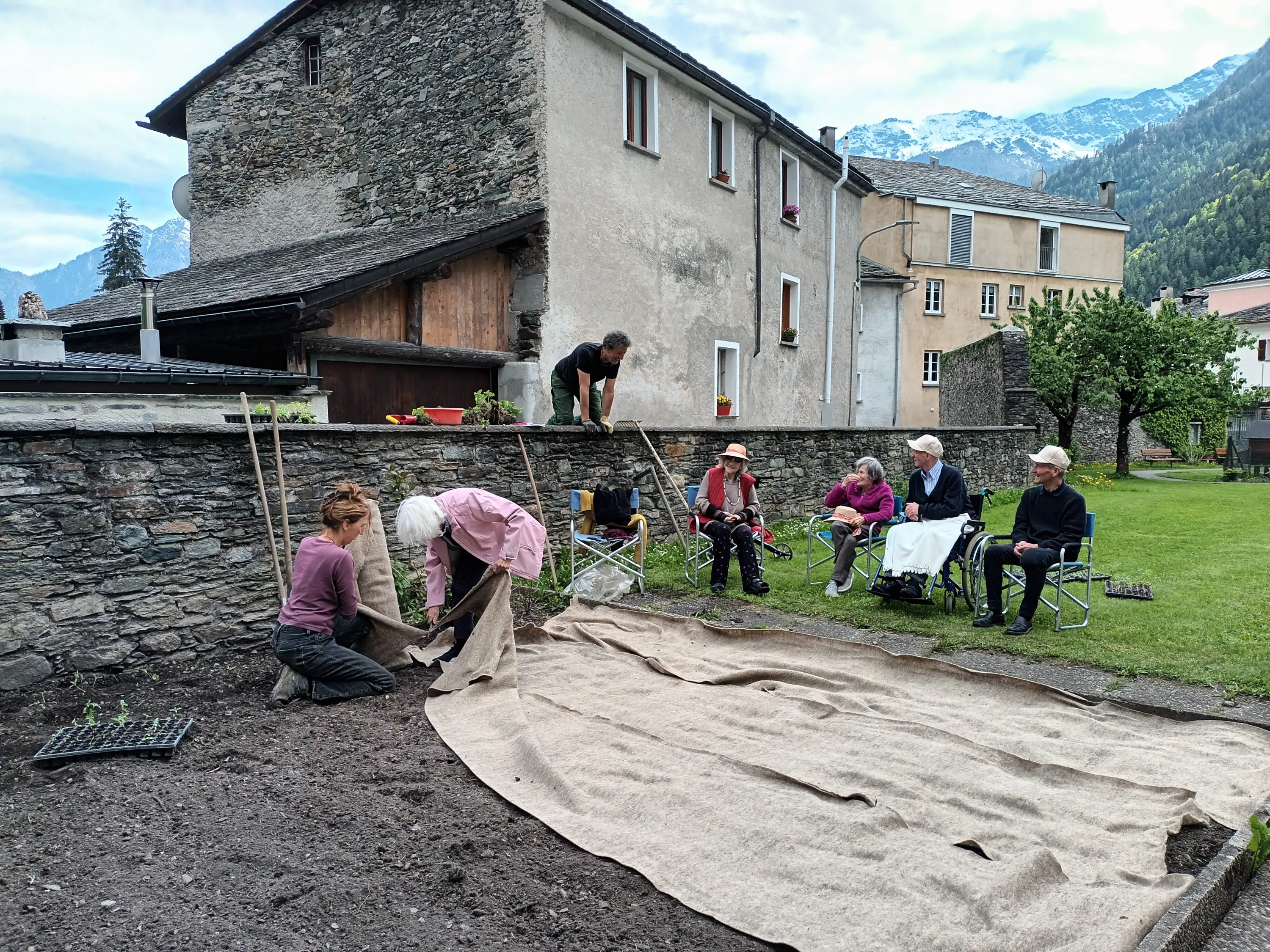 Gruppo di persone che lavorano in un orto. Alcuni sono inginocchiati a piantare, mentre altri siedono su sedie da campeggio. Sullo sfondo, si vedono edifici e montagne. Il terreno è coperto da un tessuto, e ci