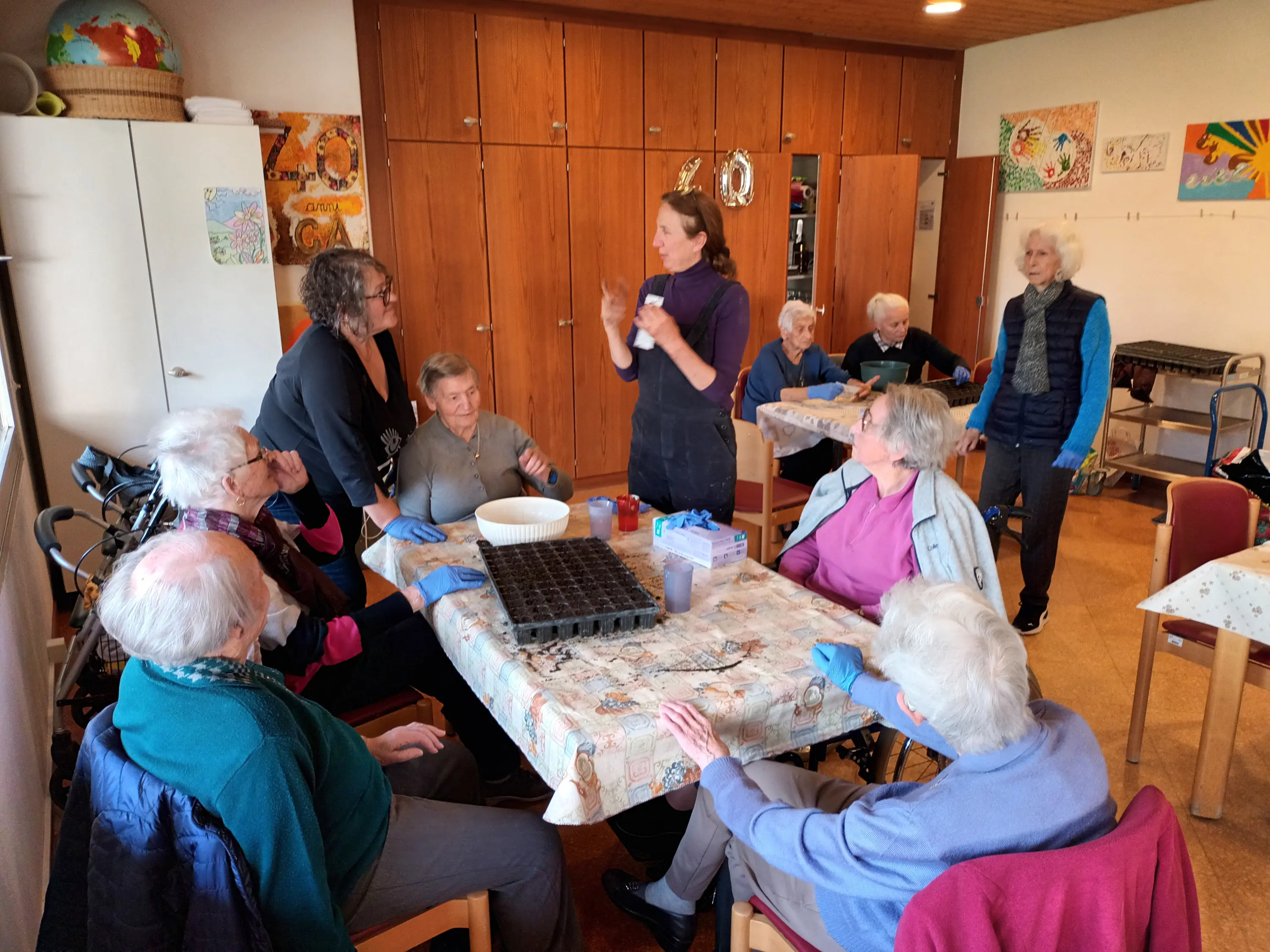 Un gruppo di anziani seduti attorno a un tavolo, mentre un'istruttrice spiega un'attività. Alcuni partecipanti osservano attentamente, altri sembrano conversare tra loro. Sul tavolo ci sono vasi e materiale per il giardinaggio. L