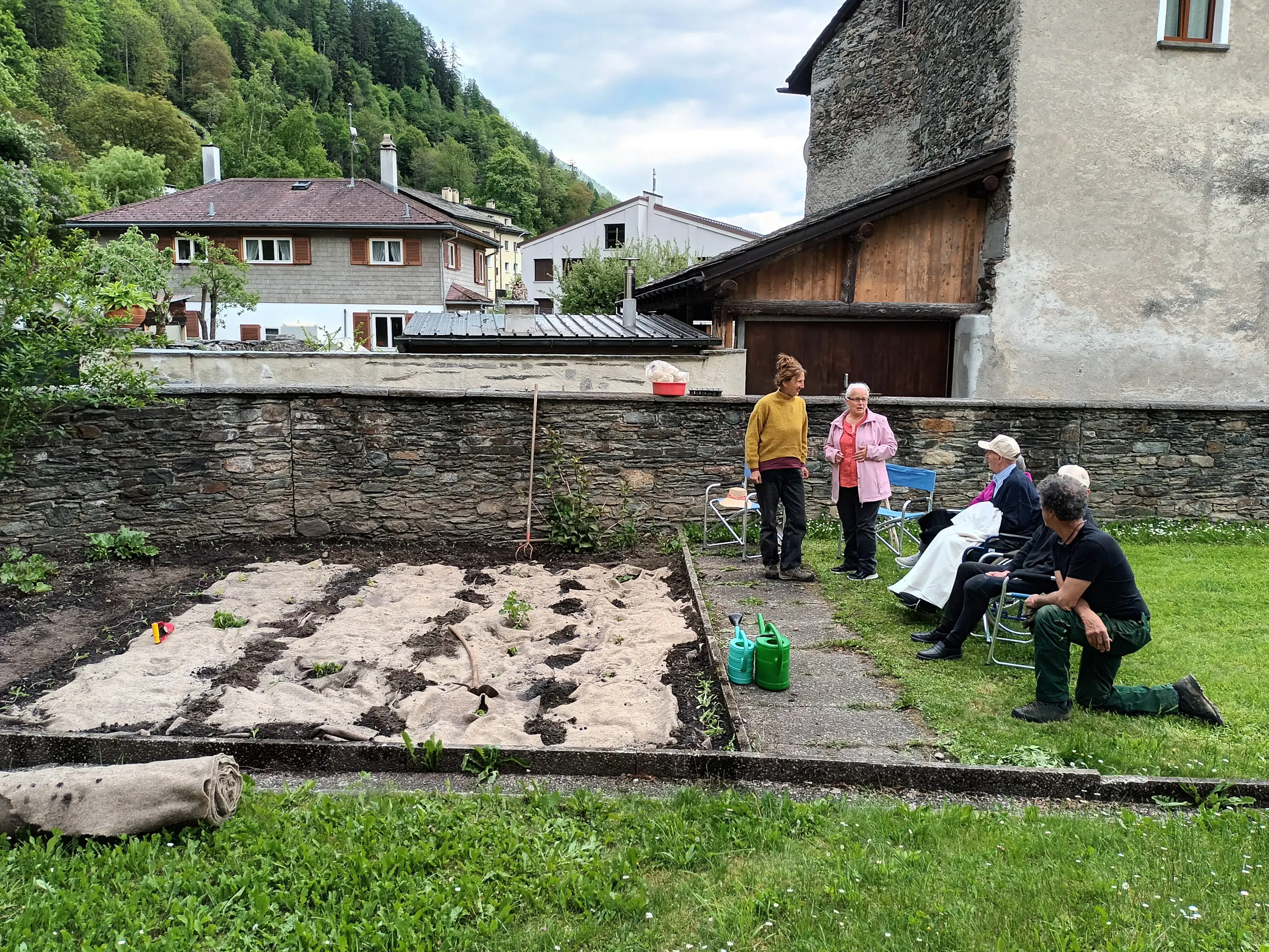 Un gruppo di persone tra cui un uomo e tre donne, discute in un giardino con un orto in un'area rurale, circondati da edifici residenziali e vegetazione. Uno dei partecipanti tiene un secchio e altri strumenti da giardinaggio sono vis