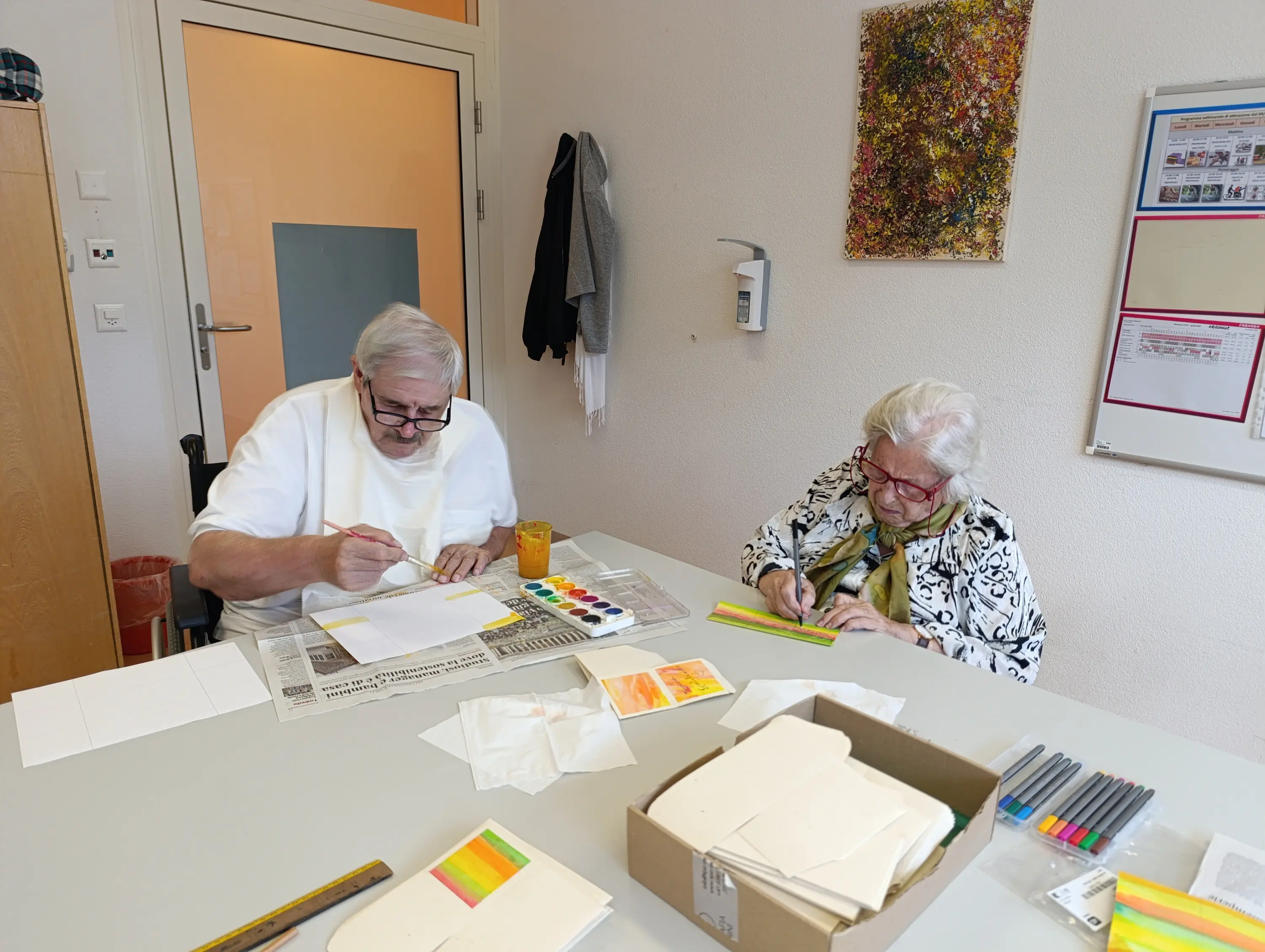 Due persone sedute a un tavolo, mentre dipingono con acquerelli. Un uomo anziano con occhiali e barba bianca usa un pennello, mentre una donna anziana con occhiali e una sciarpa verde disegna. Sul tavolo ci sono fogli