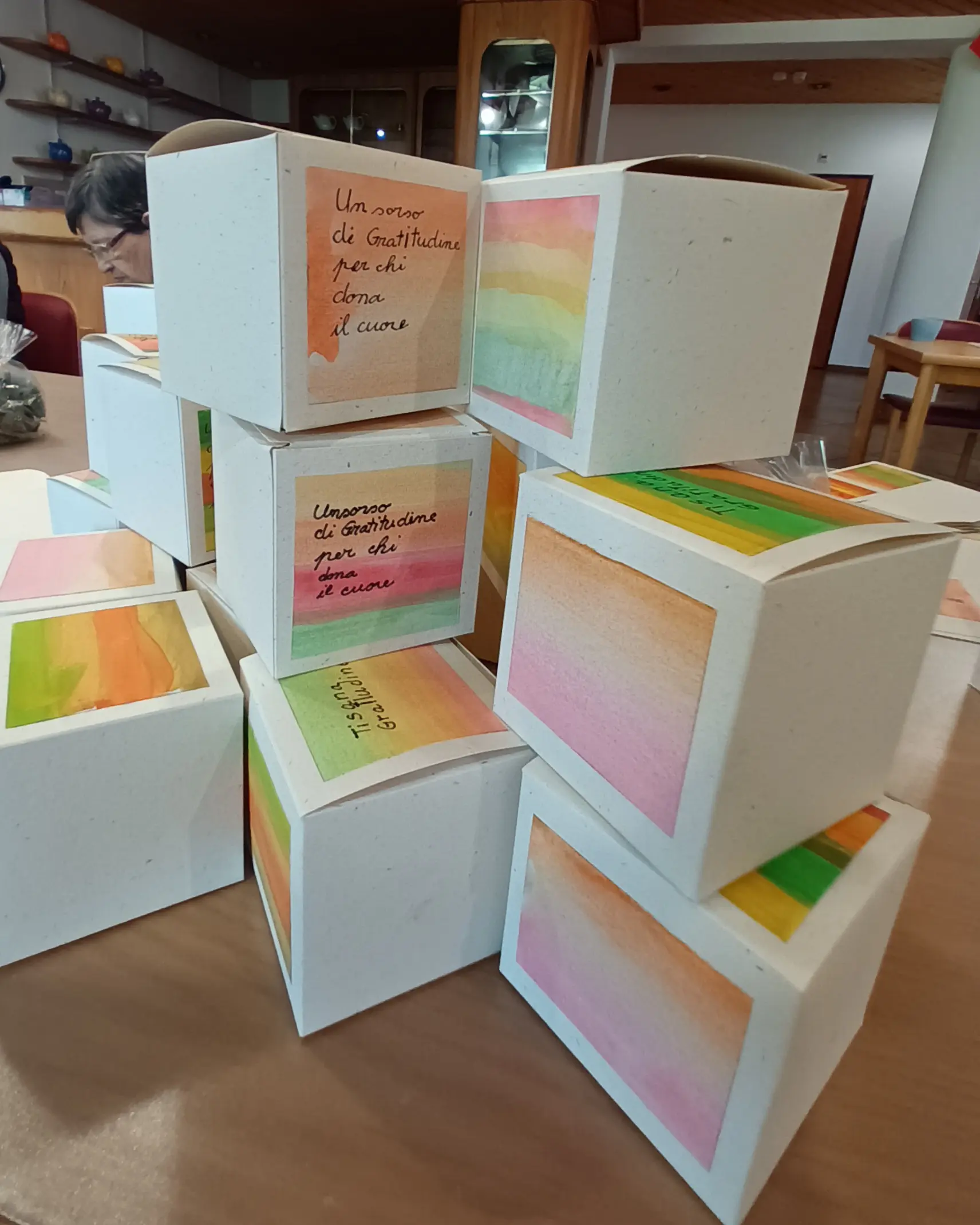 Scatole colorate impilate con sfumature di arancio, rosa e verde, su una superficie di legno. Una scatola presenta un messaggio scritto a mano che esprime gratitudine. Sullo sfondo si intravede una persona intenta a lavorare.