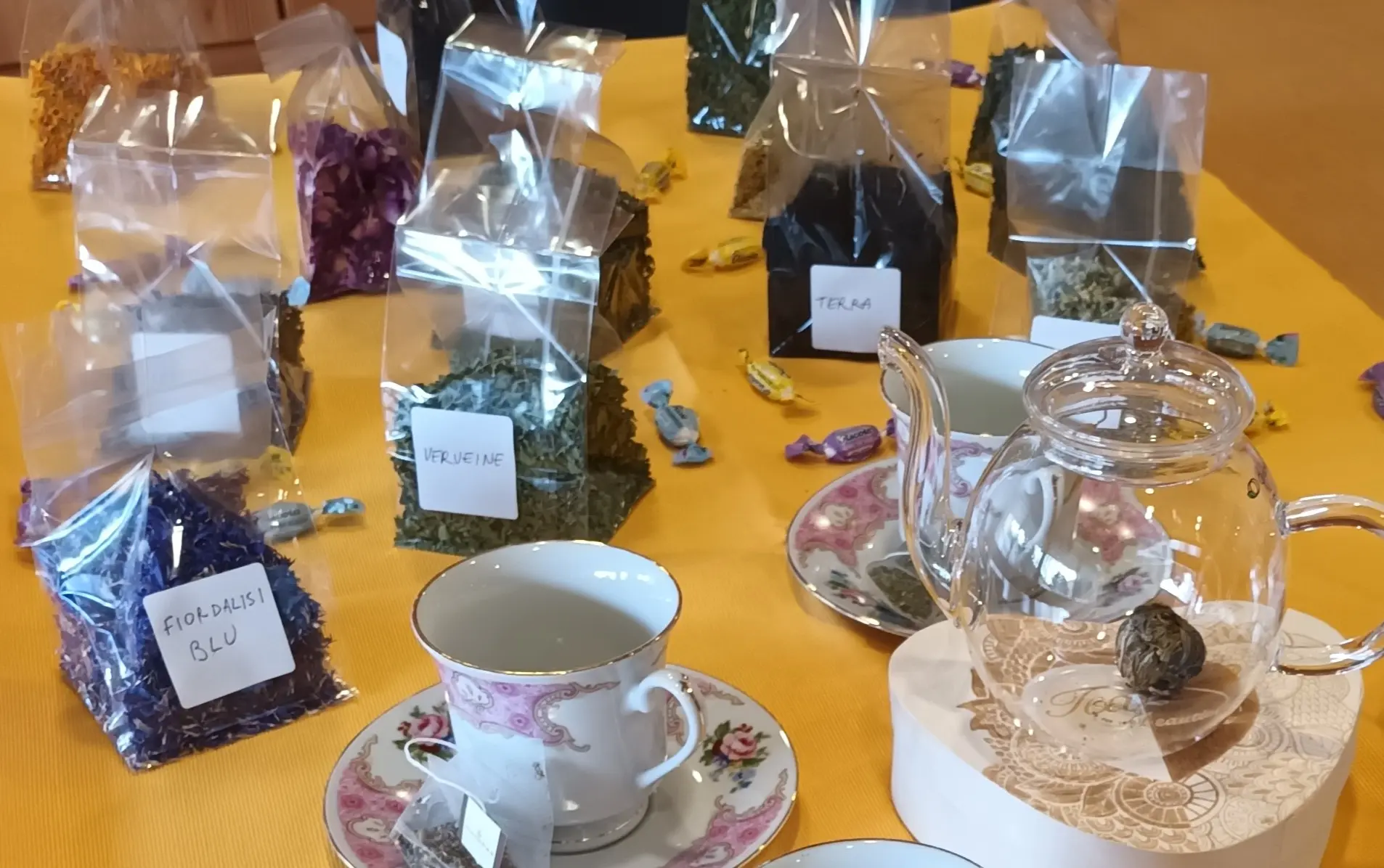 Tavolo con diverse bustine di tè e tisane colorate, alcune etichettate. Un teiera elegante e una tazza di porcellana finemente decorata sono posizionate in primo piano.