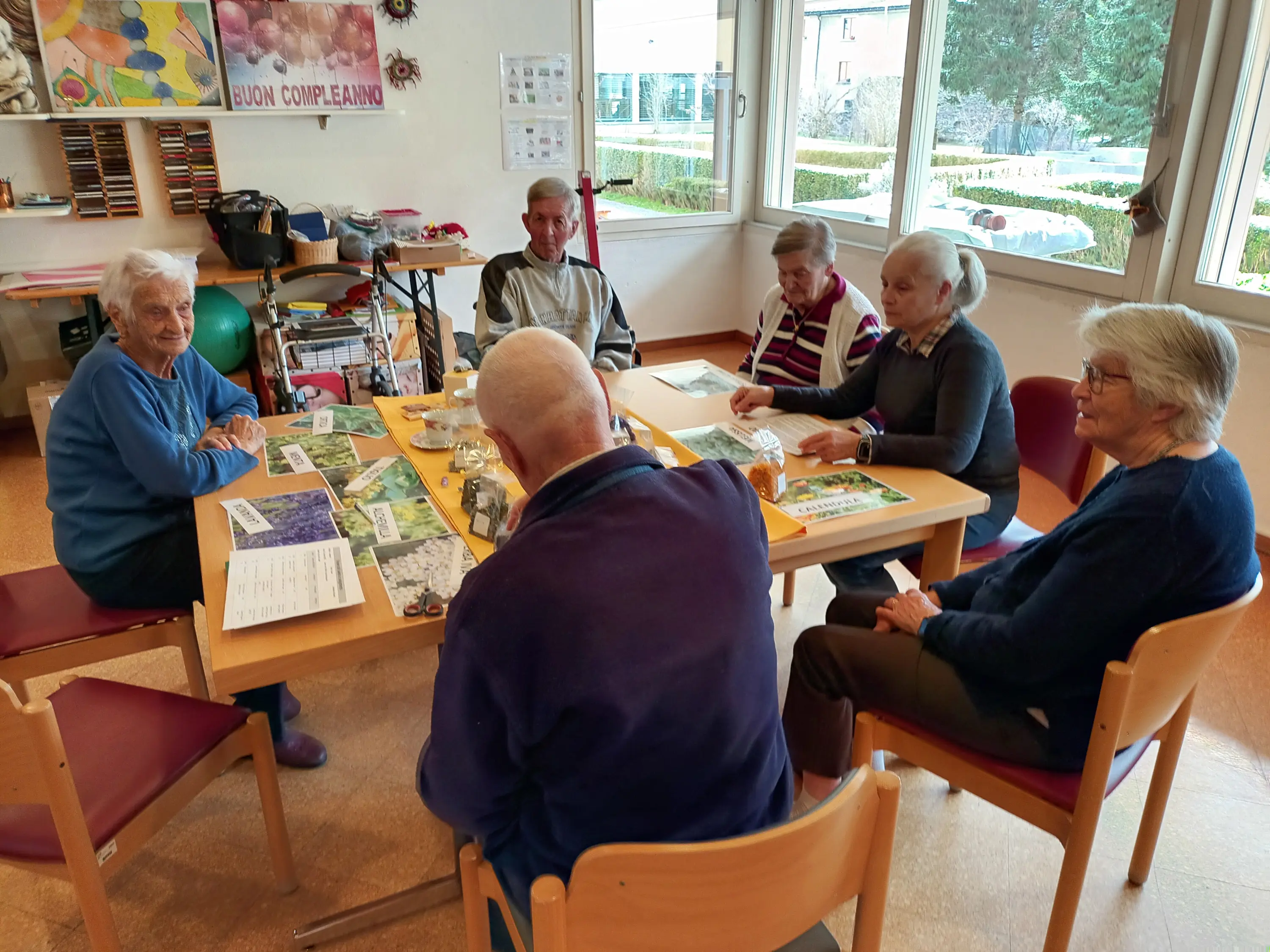Un gruppo di sei persone anziane sedute attorno a un tavolo in una sala ben illuminata. Stanno partecipando a un'attività di gruppo, con materiali di lavoro e giochi davanti a loro. Alcuni hanno espressioni sorridenti e sembrano coinvolti nella conversazione.